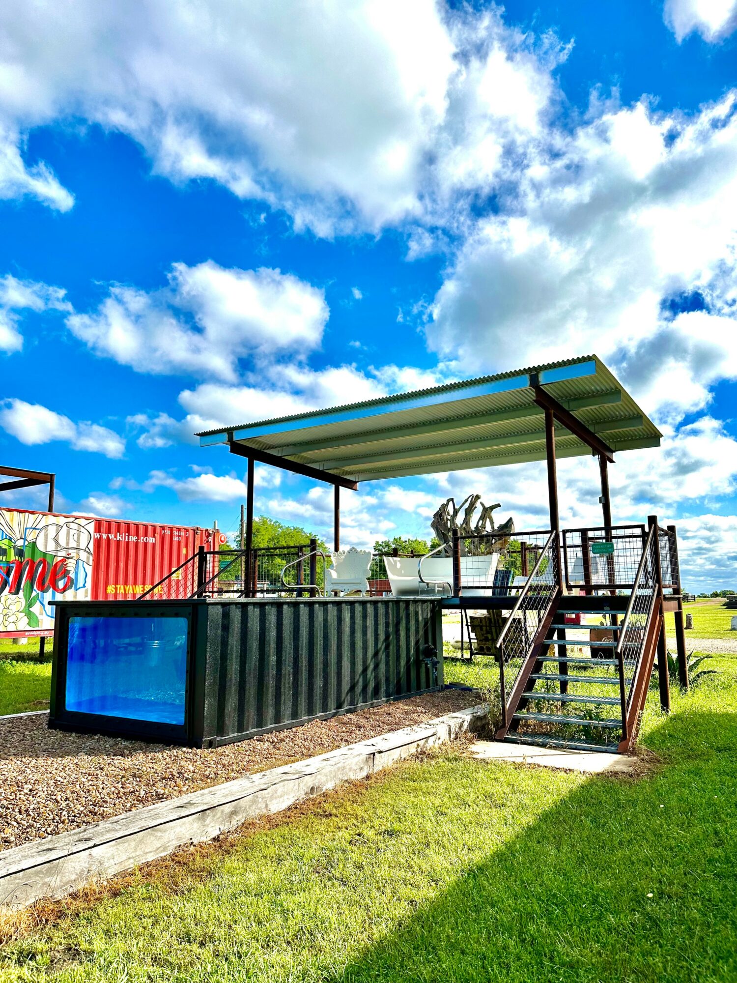 Small outdoor stage with a metal roof, stairs, and a black railing, set on grassy ground under a partly cloudy sky.