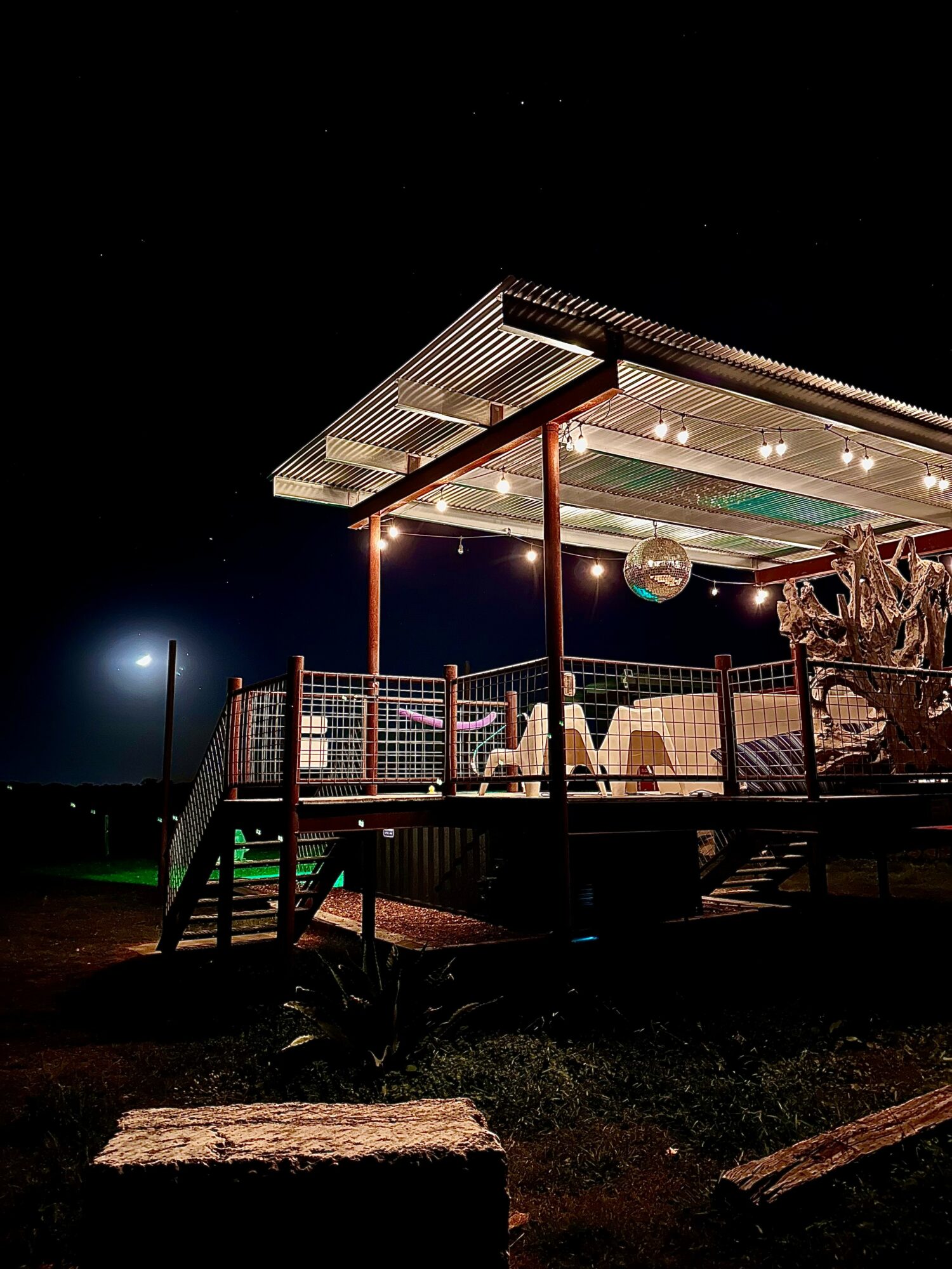 Illuminated outdoor deck with seating, a disco ball, and a tree, against a dark night sky.