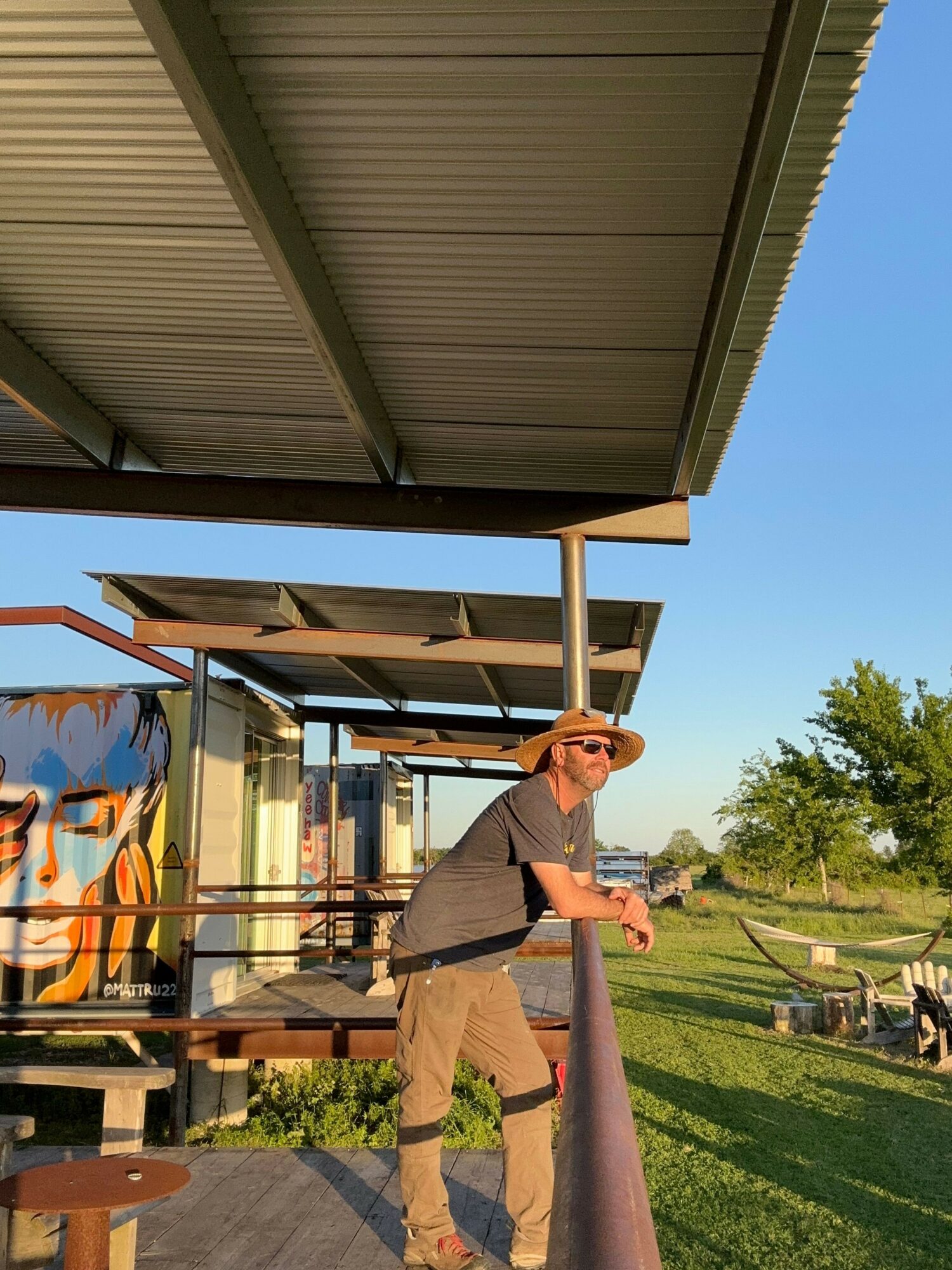Person wearing sunglasses and a hat leaning on a railing outdoors with trees and grass in background.