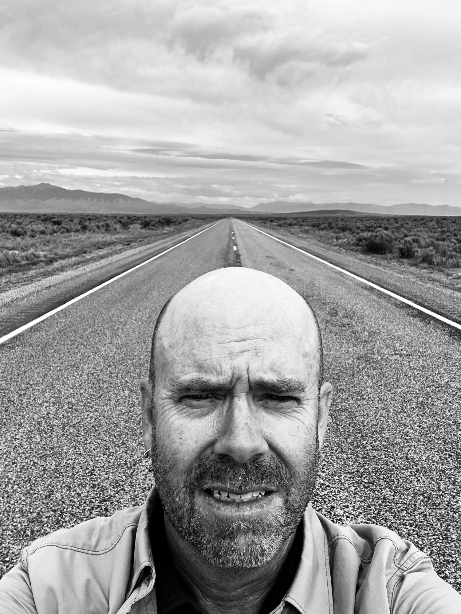 Man with a beard taking a selfie on an empty straight road in a flat landscape.