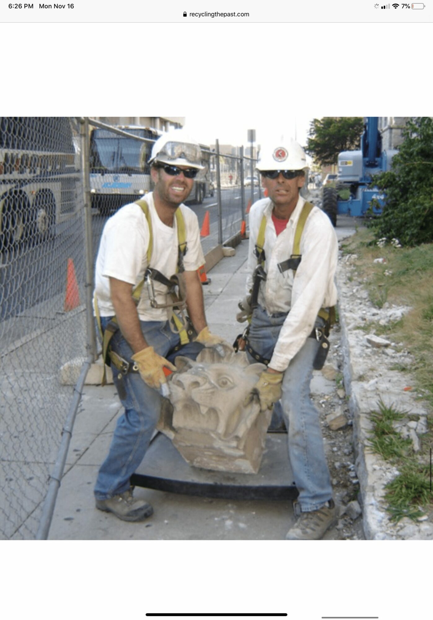 Two construction workers in safety gear holding a large stone sculpture on a cart, with a construction site background.