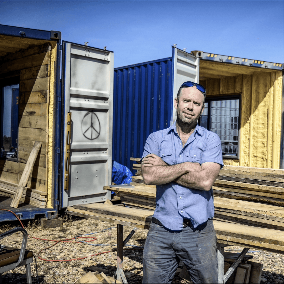 Man with crossed arms standing in front of colorful shipping containers and wooden structures.