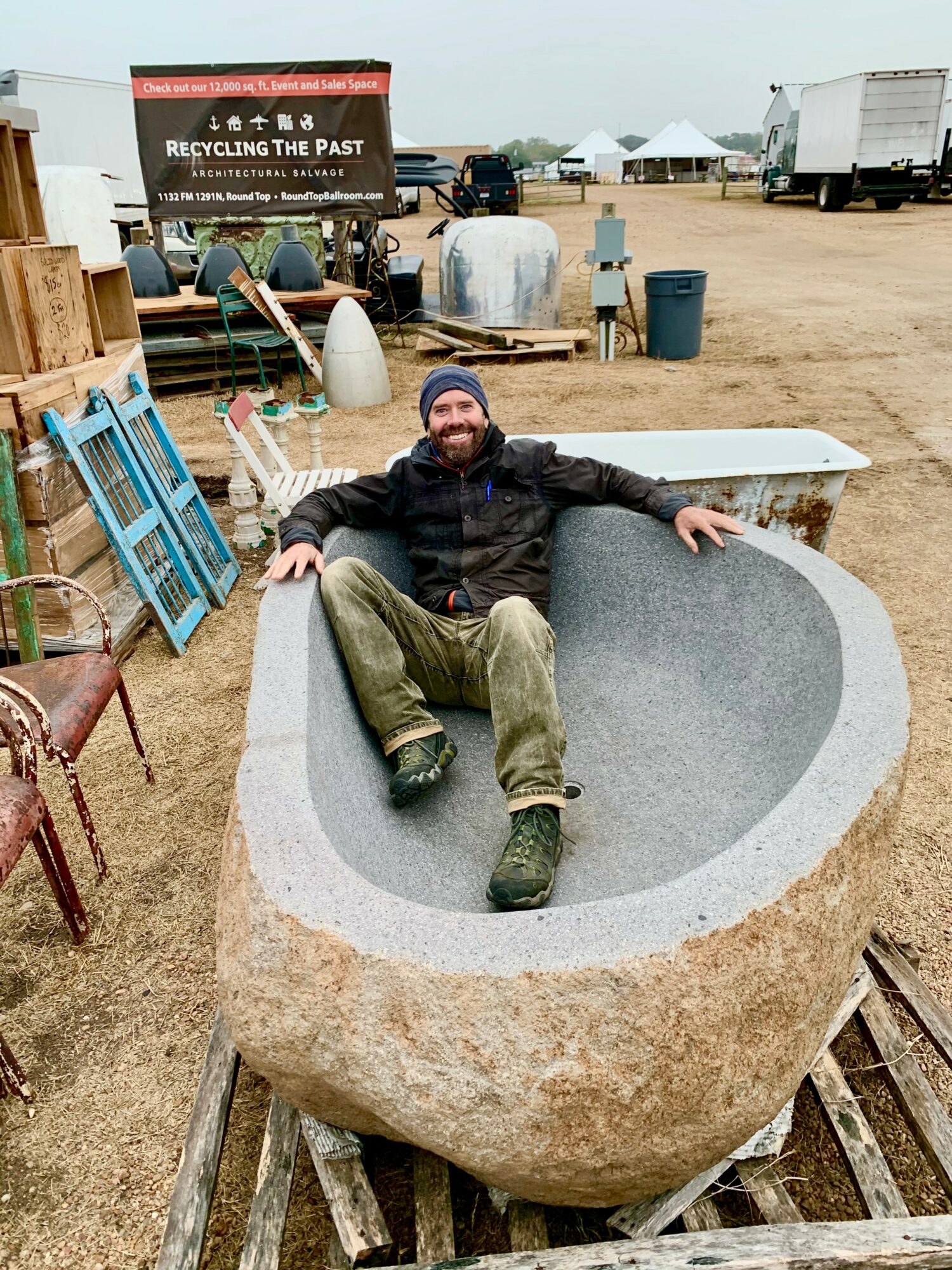 Man sitting inside a large stone bathtub at an outdoor market or event, surrounded by various objects and tents.