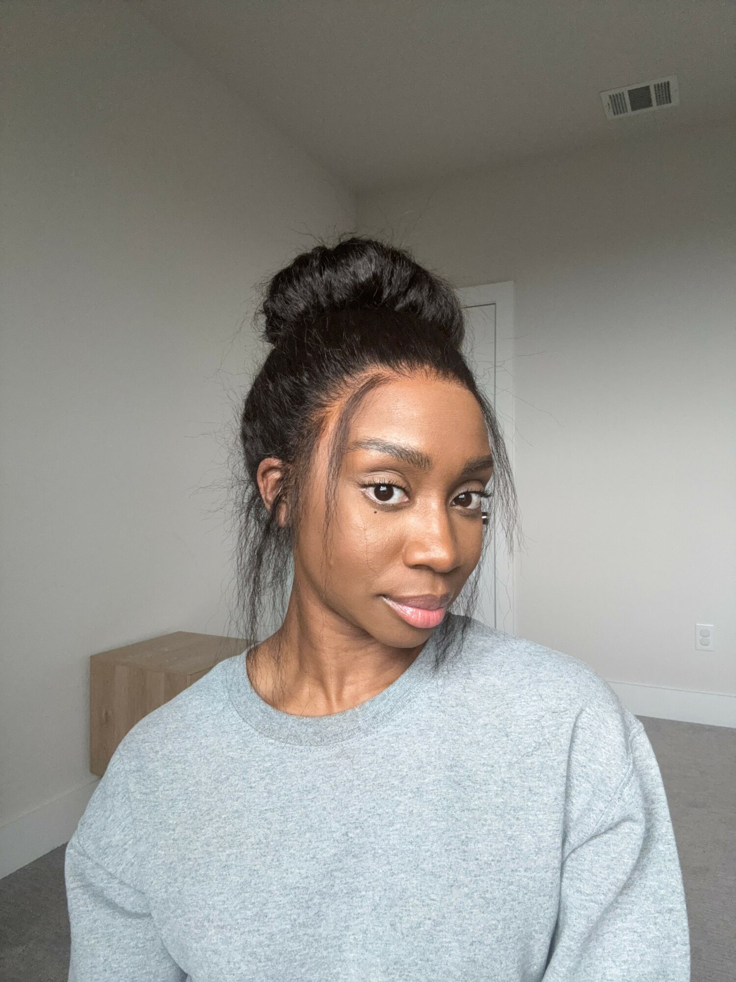 Woman with dark hair in a bun, wearing a gray shirt, looking to the side in a room with white walls.