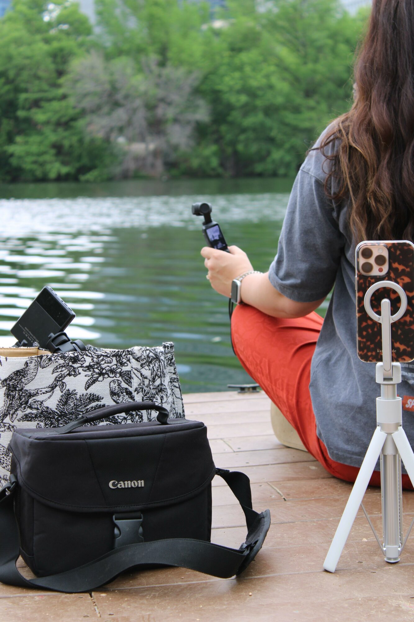 Person sitting by water holding a remote control, with camera bag and equipment nearby, outdoors with trees and water.