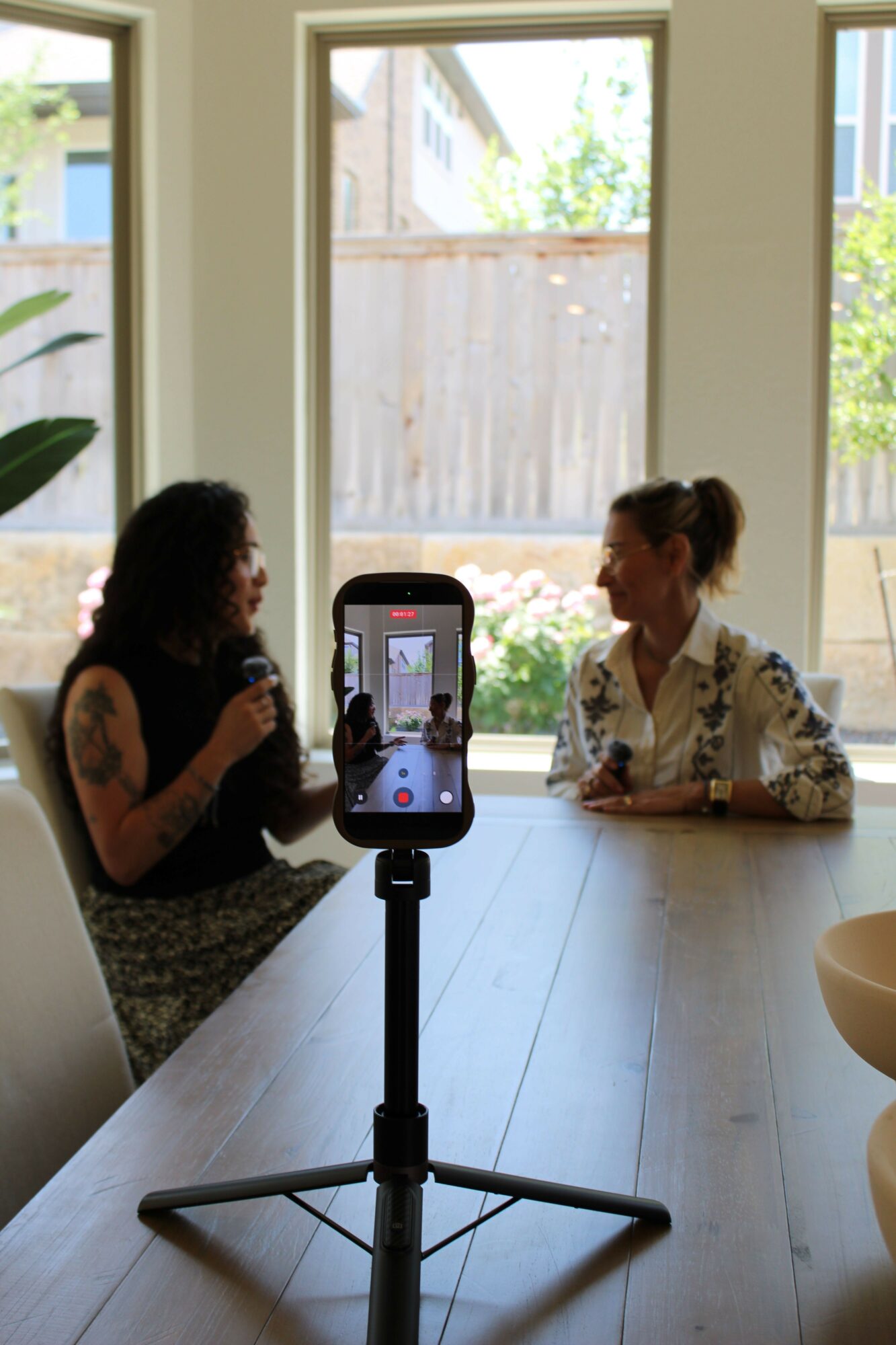 Two women sit at a table, recording a video with a smartphone on a tripod, in a bright room with large windows.