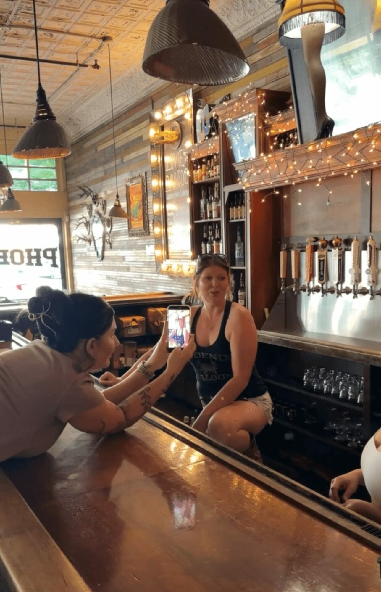 Person taking a photo of a woman sitting at a bar counter inside a pub or bar with warm lighting and wooden decor.