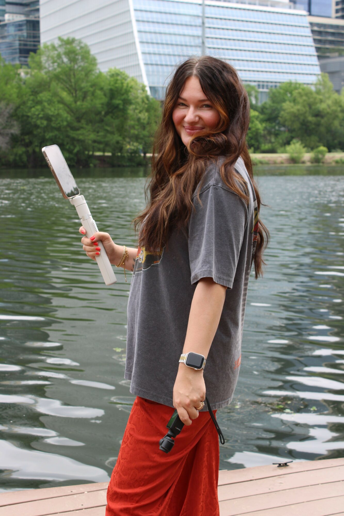 Young woman with long hair smiling, holding a tennis racket, standing near water with trees and modern buildings in background.