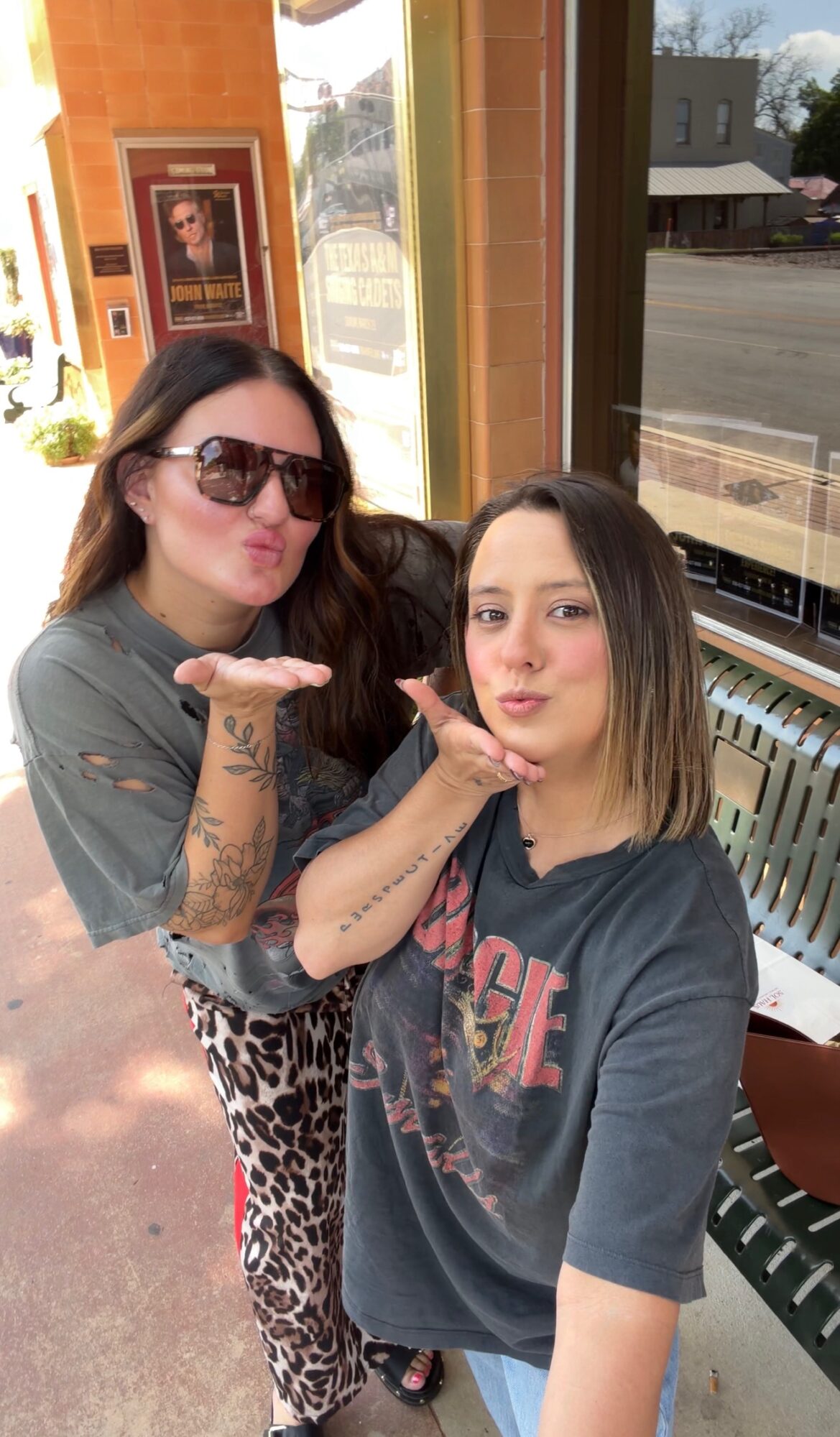 Two women posing indoors, one with sunglasses and tattoos, the other with short hair, both making kiss gestures.
