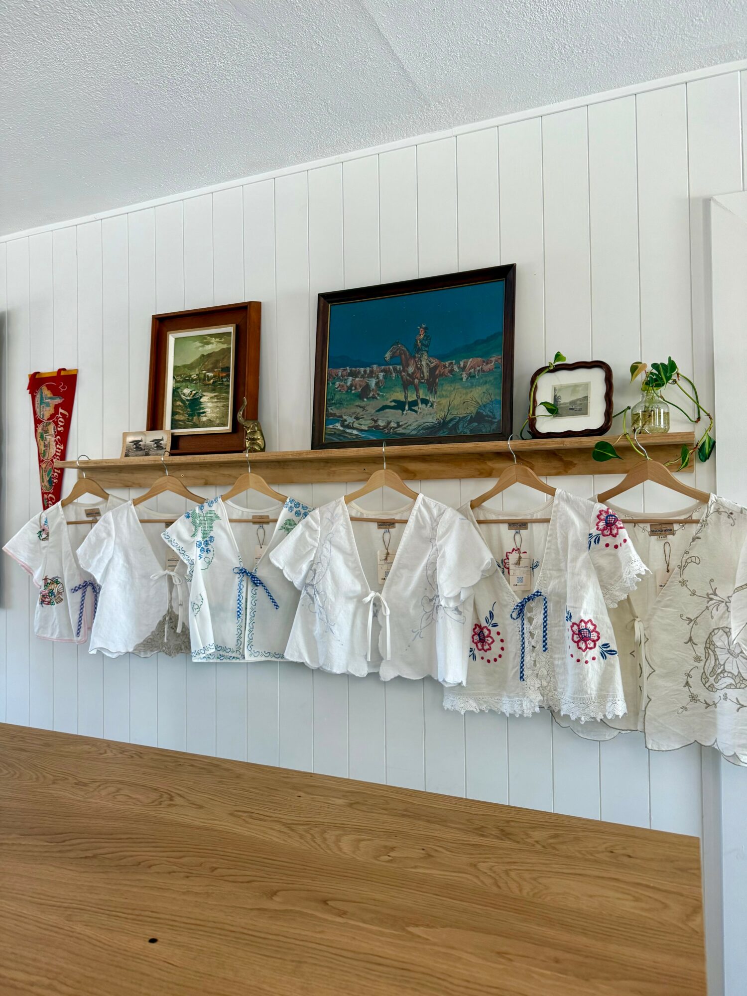 Child's white dresses hanging on a wooden rod with framed pictures and a plant on a shelf above.