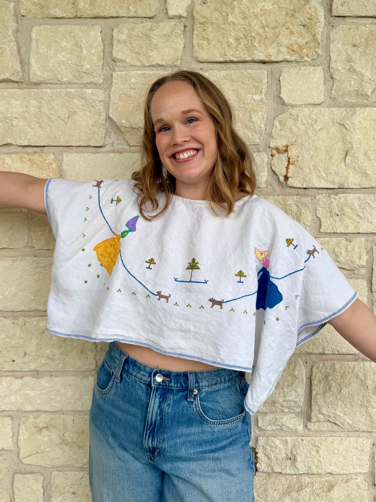 Young woman with shoulder-length wavy hair smiling, wearing a white embroidered top and blue jeans, standing against a stone wall.