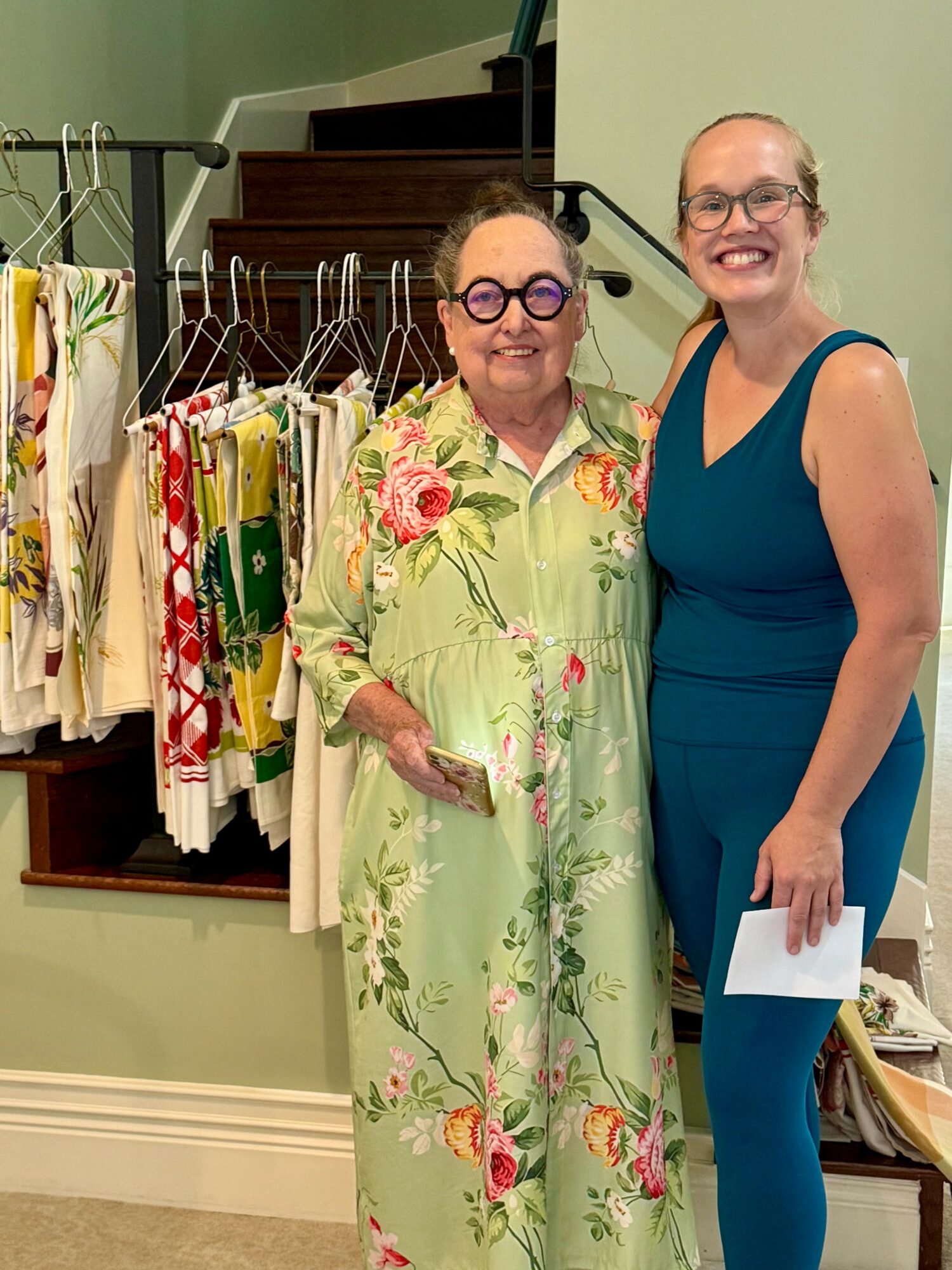 Two women standing indoors, smiling, with clothing racks behind them displaying colorful garments.