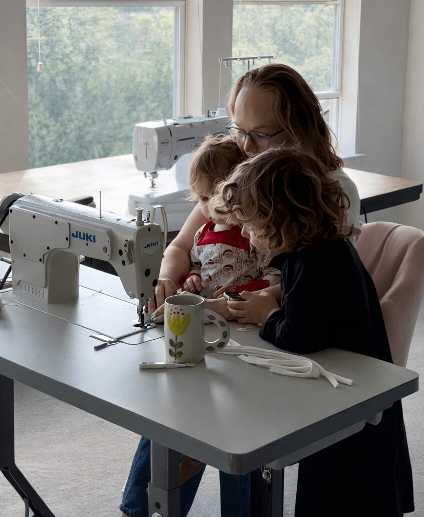 Woman with two children sitting at a table with sewing machines, cups, and utensils near a window.