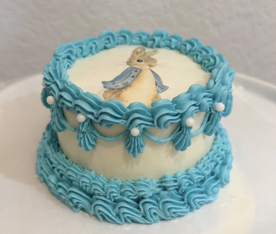 Round cake with blue piped borders and a bunny illustration on top, decorated with white pearls.