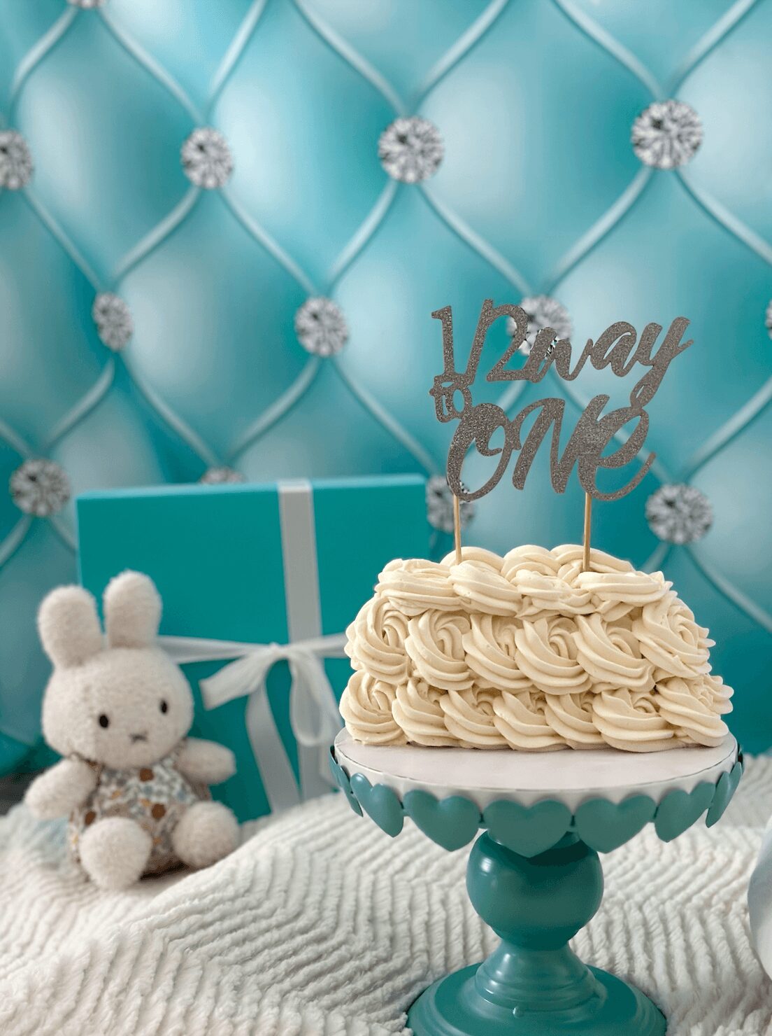 Cream-colored cake with piped frosting on a teal cake stand, decorated with a cake topper reading '12 and Me'.