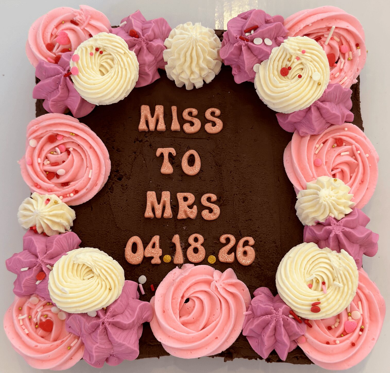 Square chocolate cake with pink and white frosting flowers, decorated with message and date in the center.