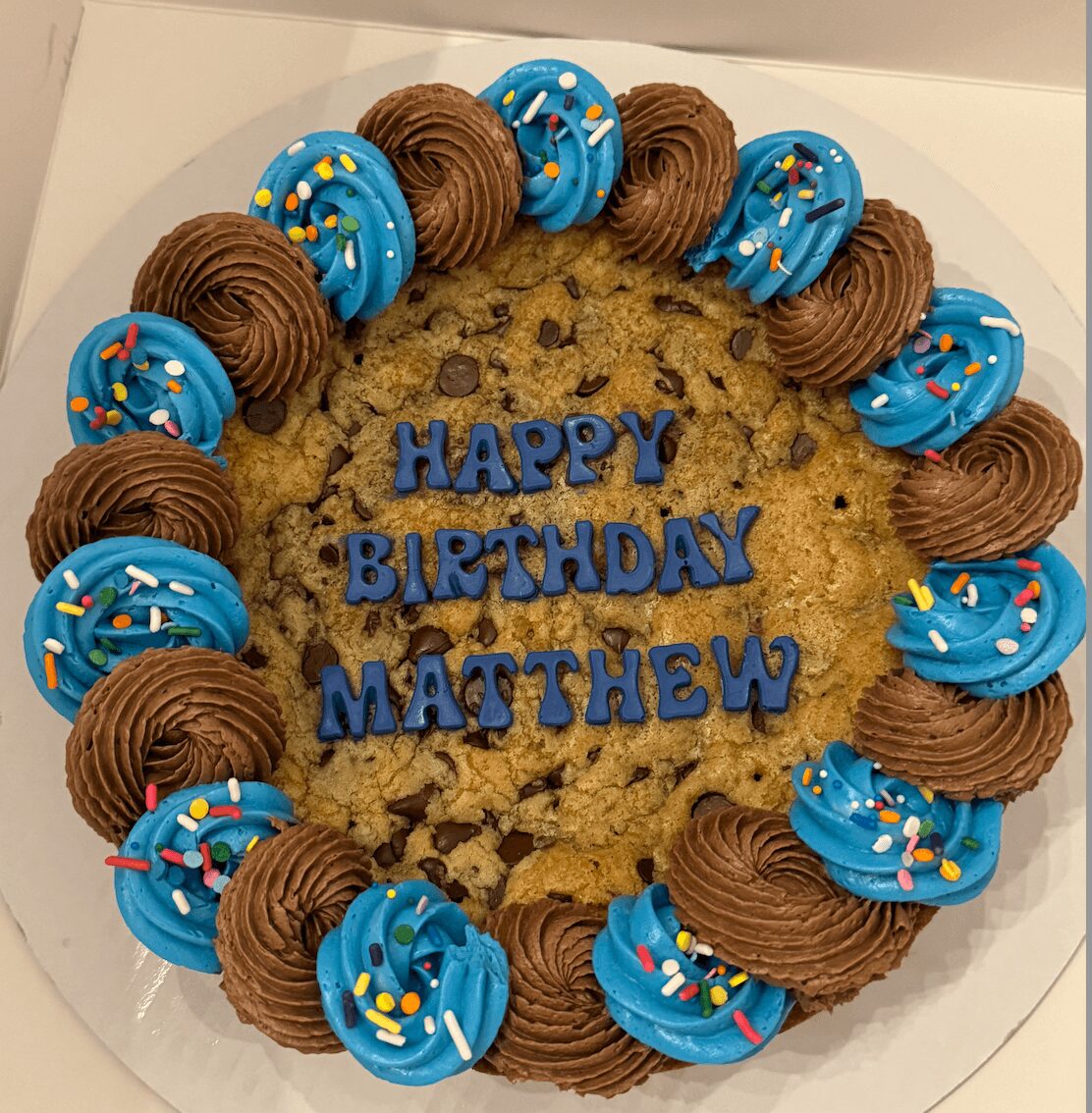 Chocolate chip cookie with blue and brown frosting swirls and sprinkles, birthday message for Matthew, on a white surface.
