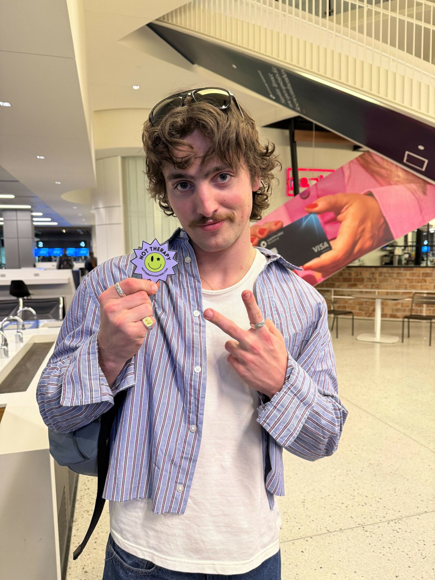 Young man with curly hair holding a smiley face badge and pointing at it, in an indoor shopping area.