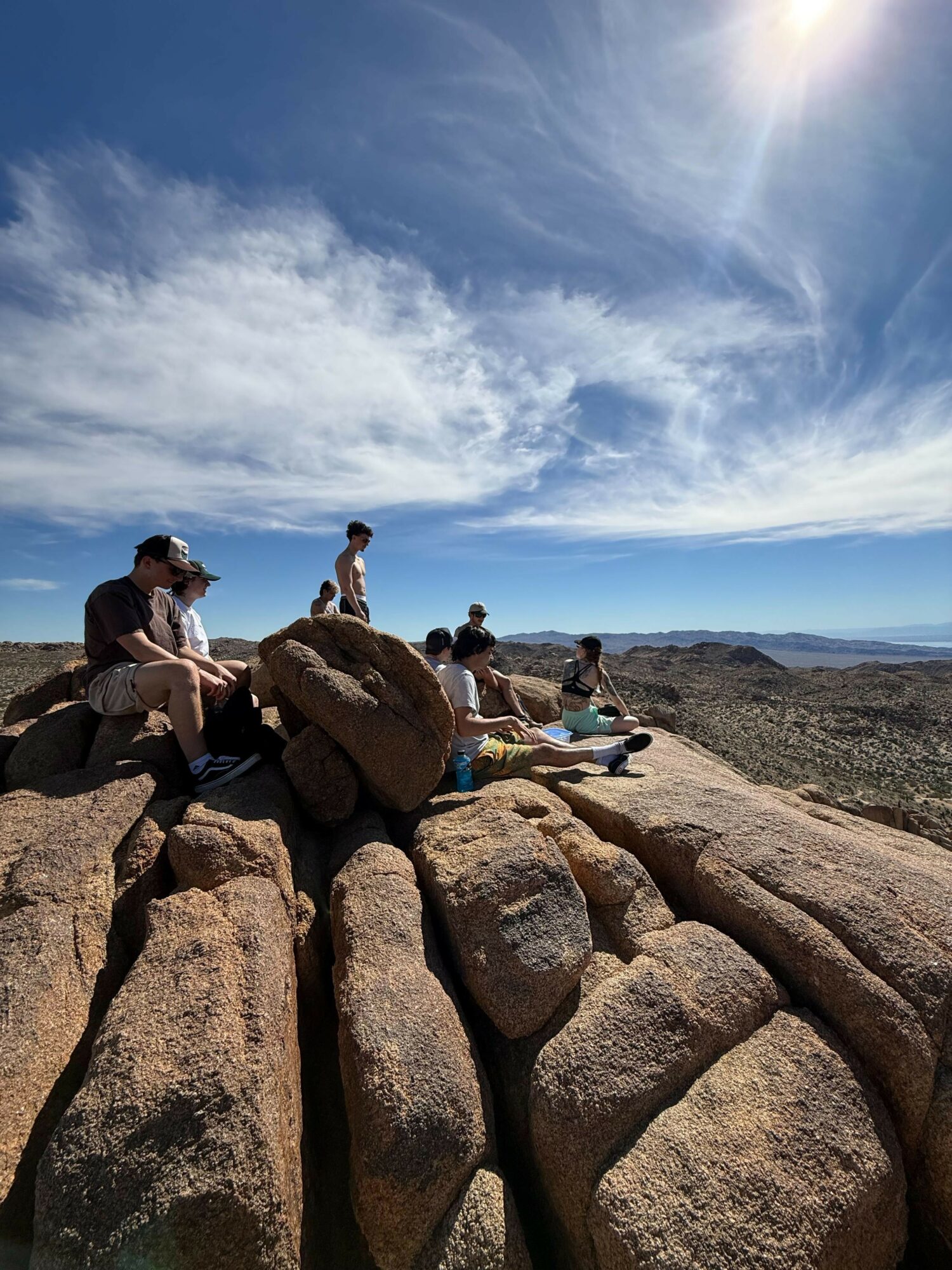 Group of people sitting on rocks outdoors under a partly cloudy sky with the sun shining.