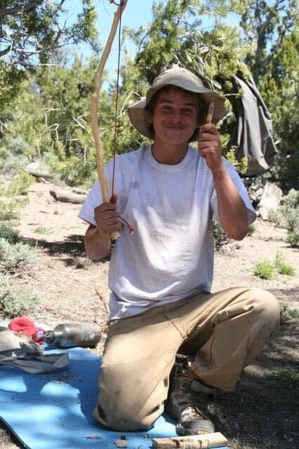 Person kneeling outdoors holding a stick, wearing a hat and casual clothing, with trees and sky in background.