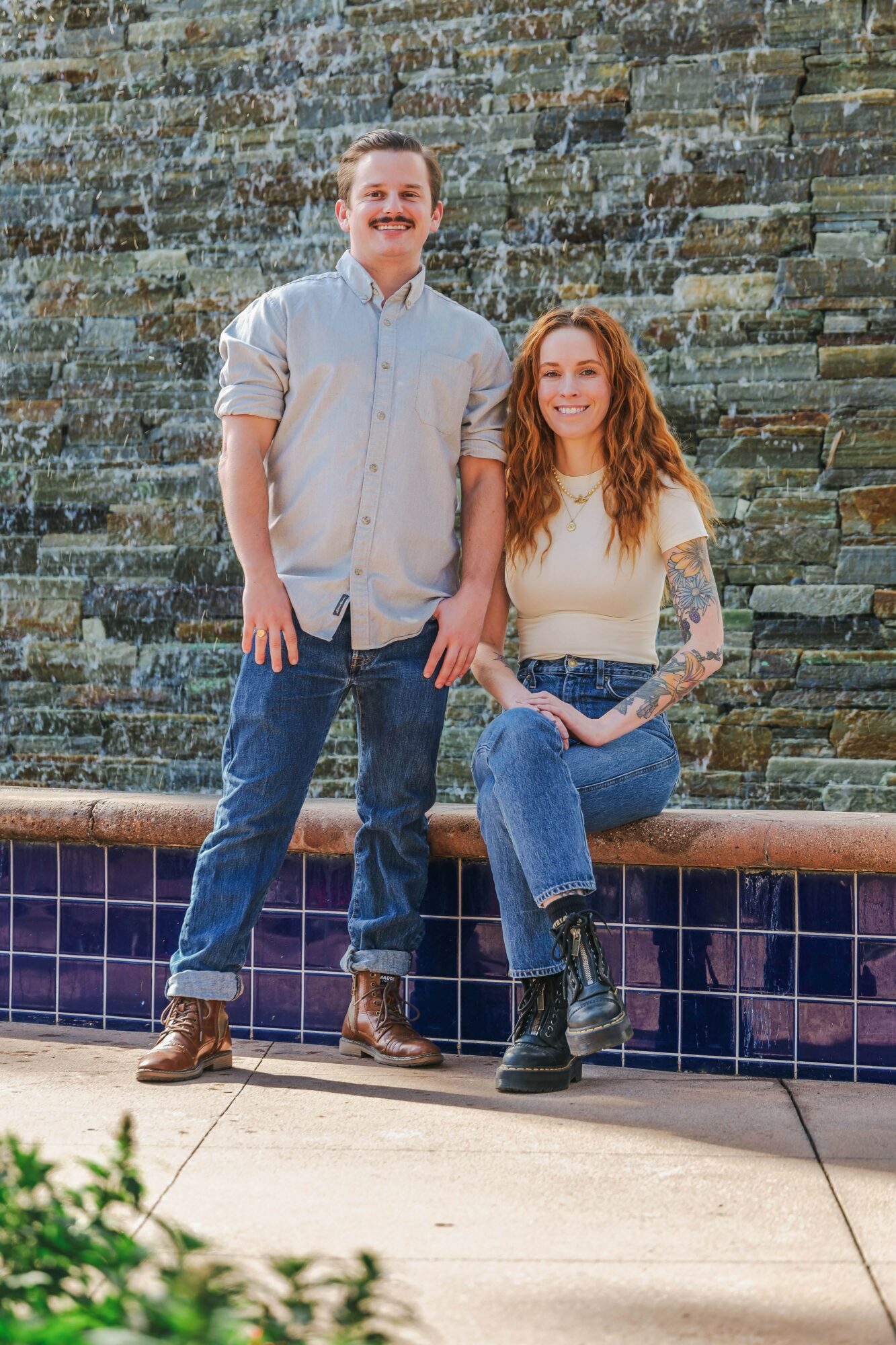 A man and woman stand in front of a stone wall, smiling, with the woman sitting on a ledge.