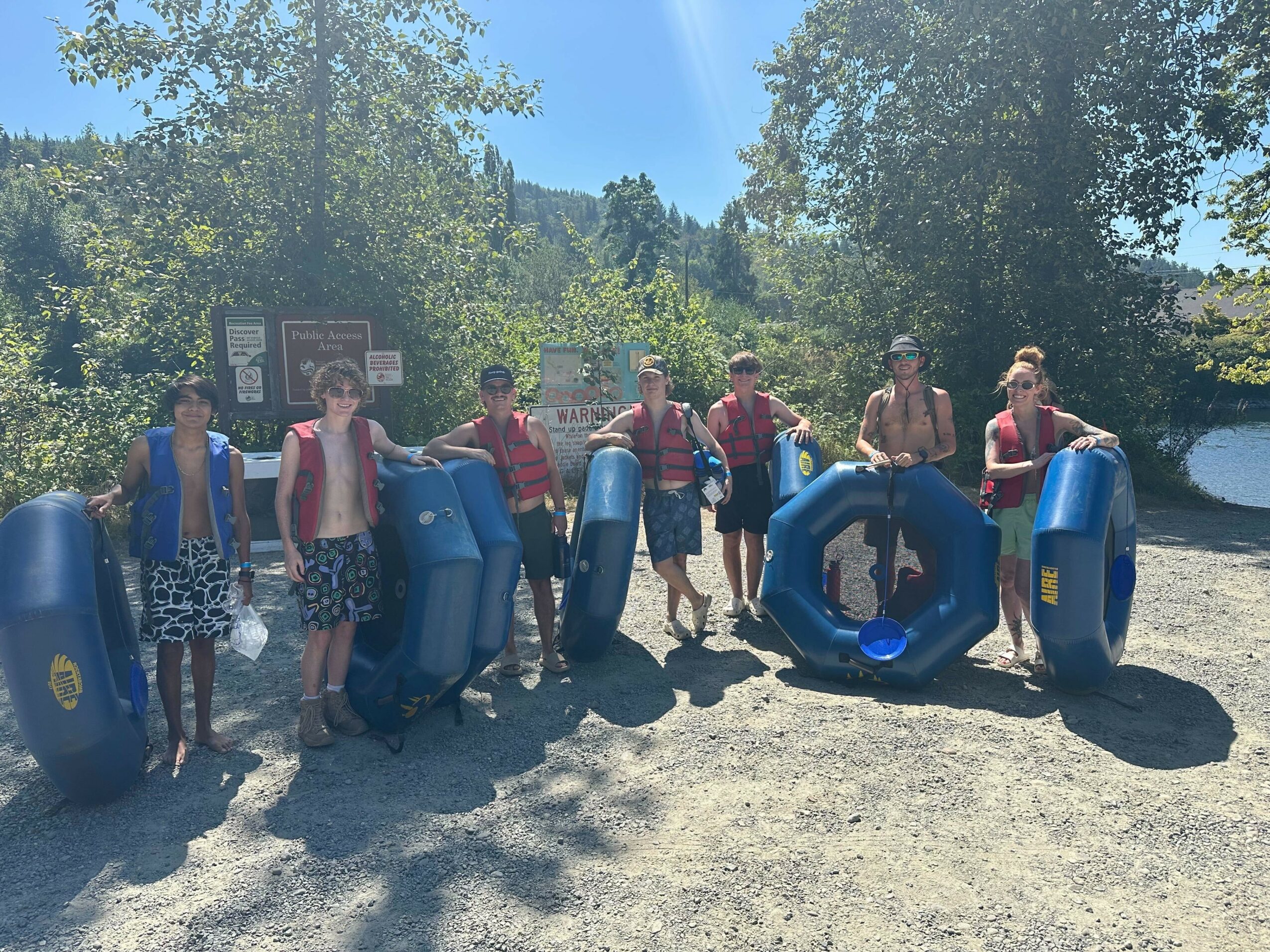 Group of seven people holding blue inner tubes outdoors on a sunny day, trees and a river in background.