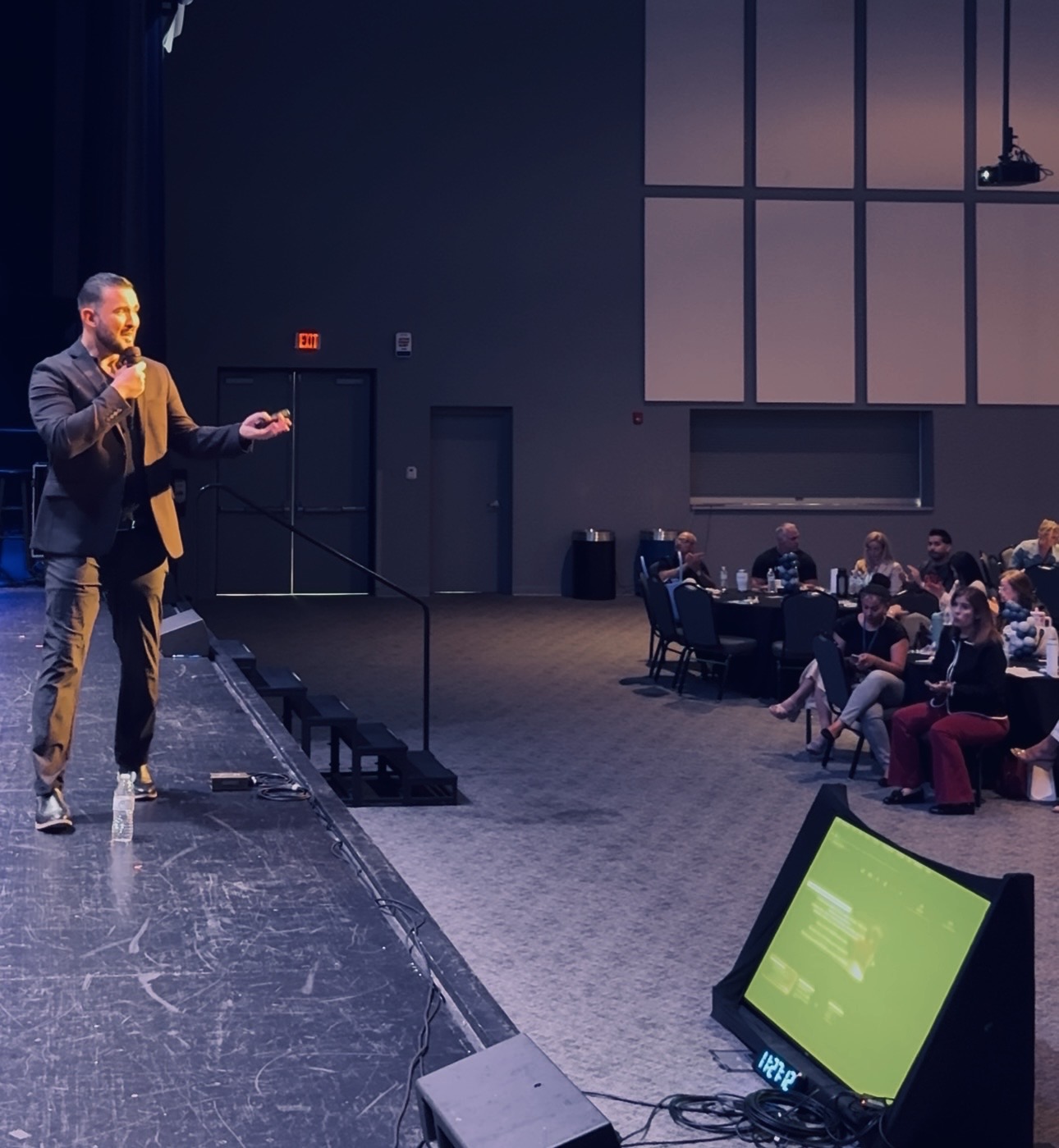 Man speaking on stage to audience in a conference room with a large screen, audience seated at tables.