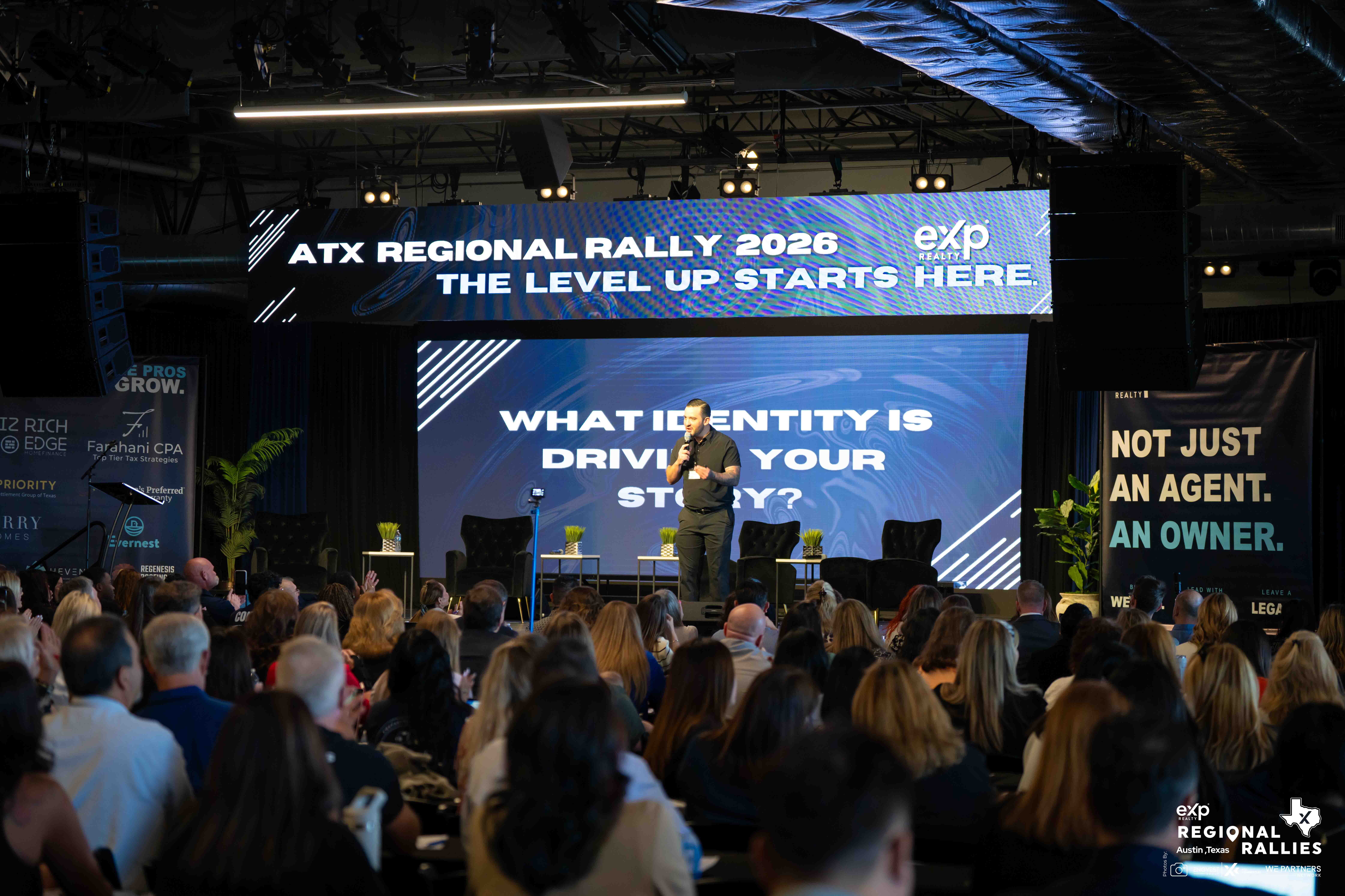Speaker on stage at rally with audience in foreground, large screen displays text about identity and story, event setting indoors.