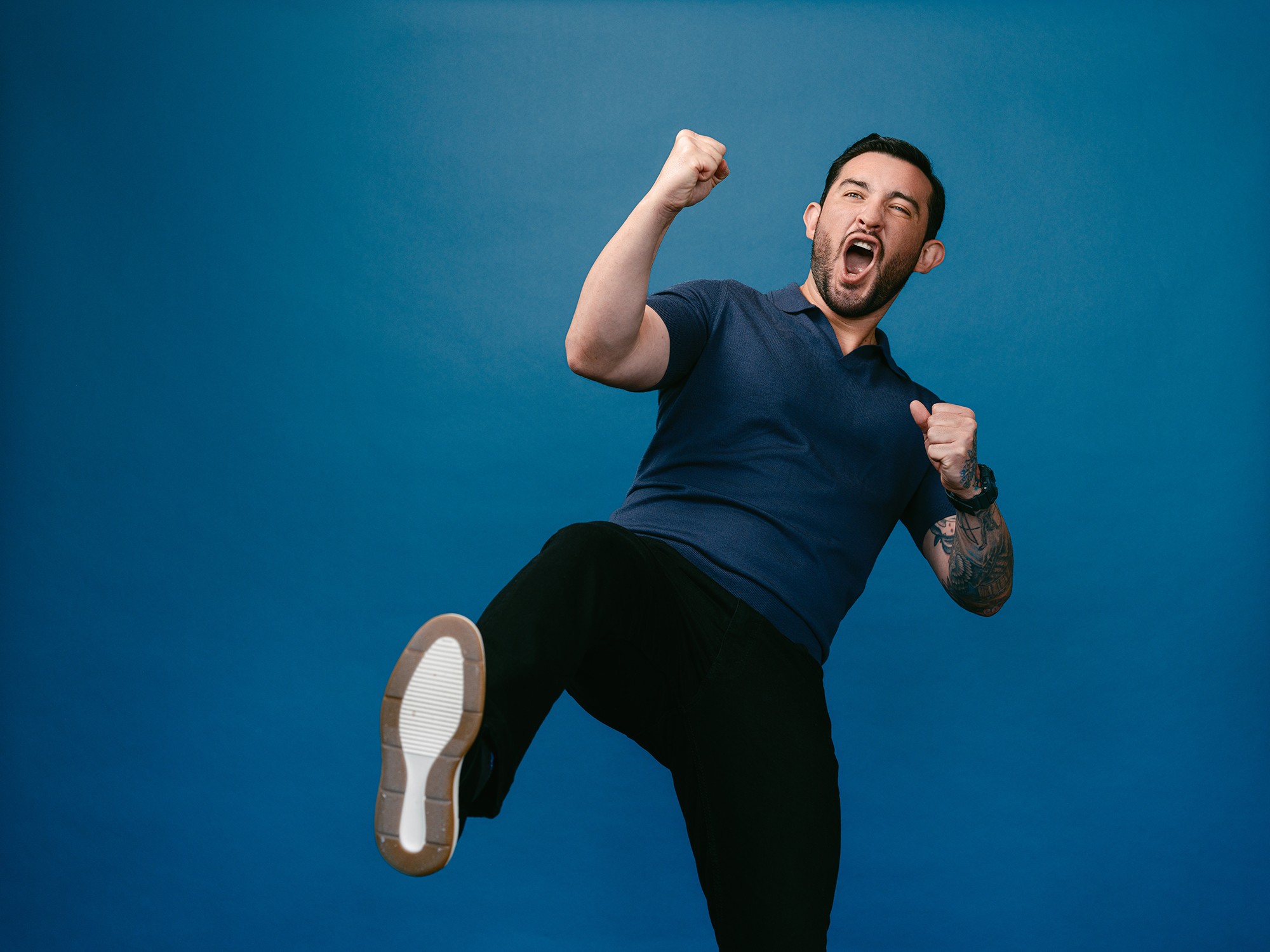 Man celebrating with raised fist and open mouth, wearing a navy shirt and black pants, against a blue background.