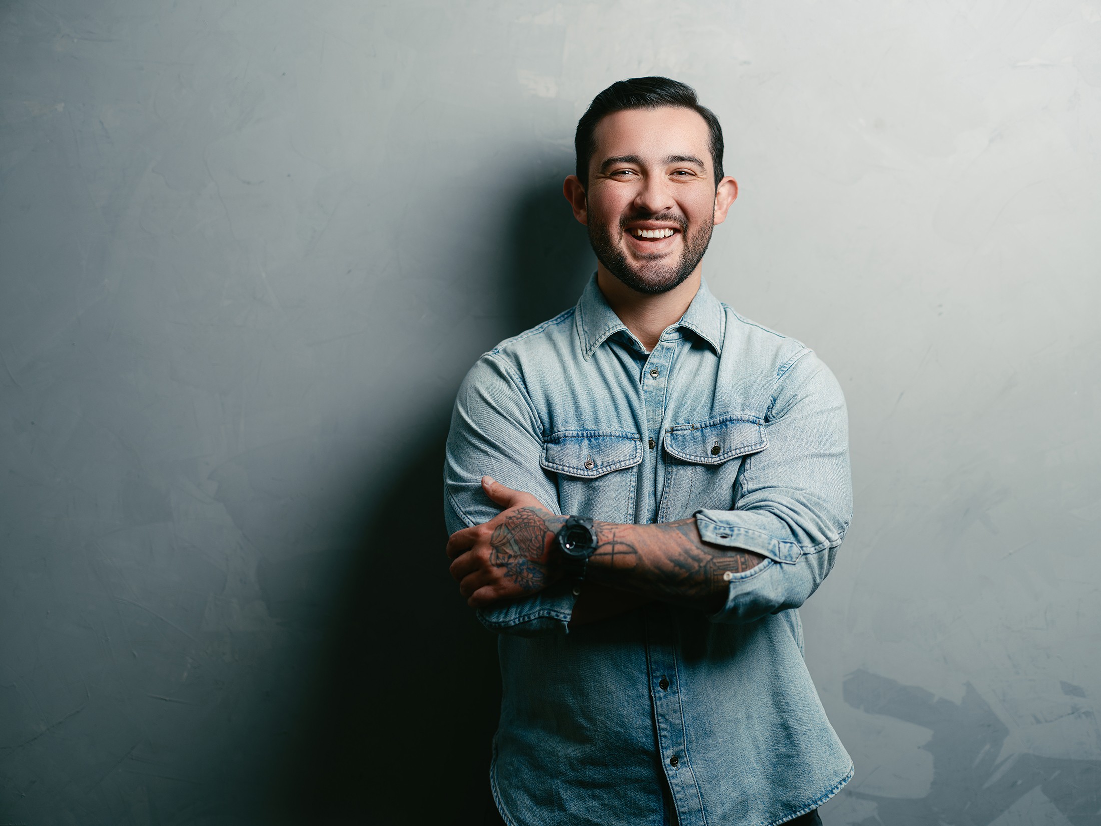 Smiling man with dark hair and beard, wearing a light denim shirt, standing against a plain gray wall.