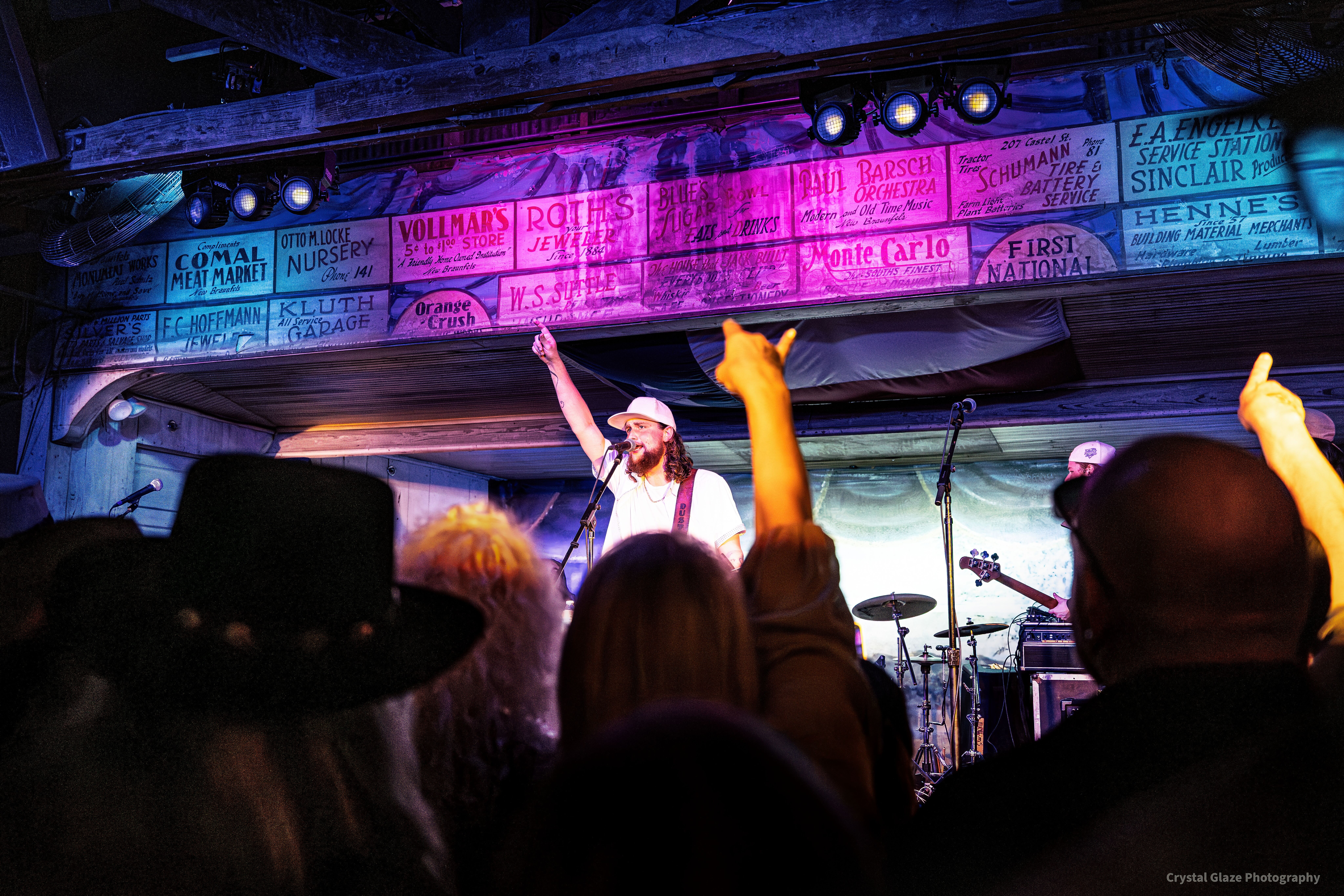 Performer on stage with microphone, audience raising hands, colorful banners above, dim lighting, lively atmosphere.