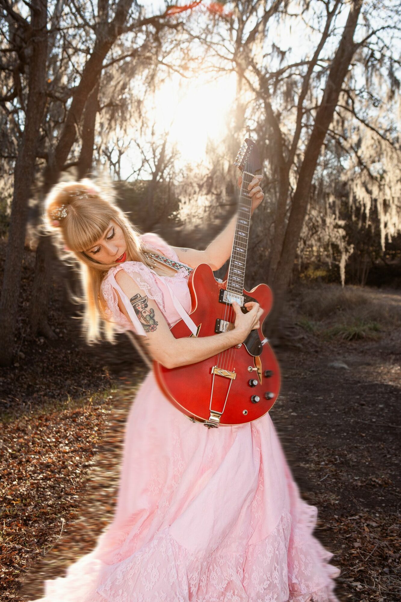Young woman with blonde hair in a pink dress holding a red electric guitar outdoors among trees.