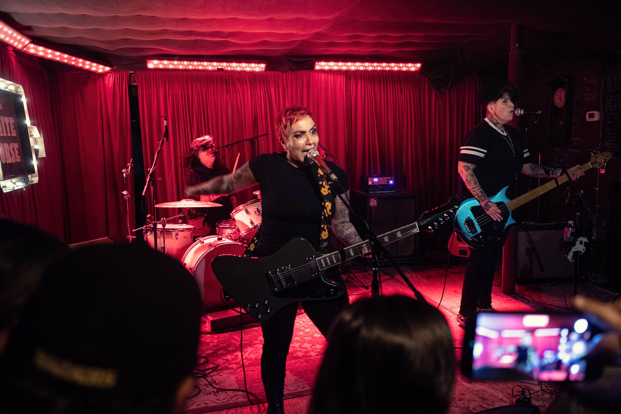 Band performs on stage with red curtains, singer with guitar, drummer, and bassist, audience members visible in foreground.