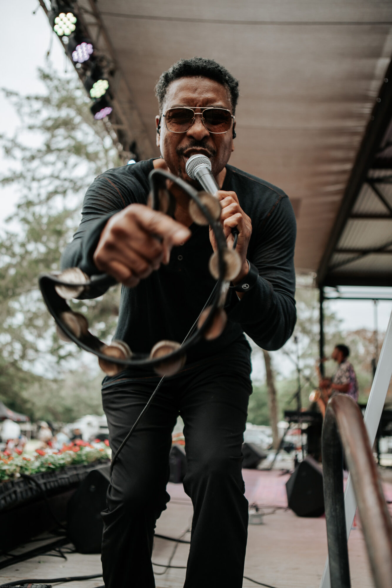 Man singing into microphone, holding a tambourine, wearing sunglasses, outdoors stage setting.