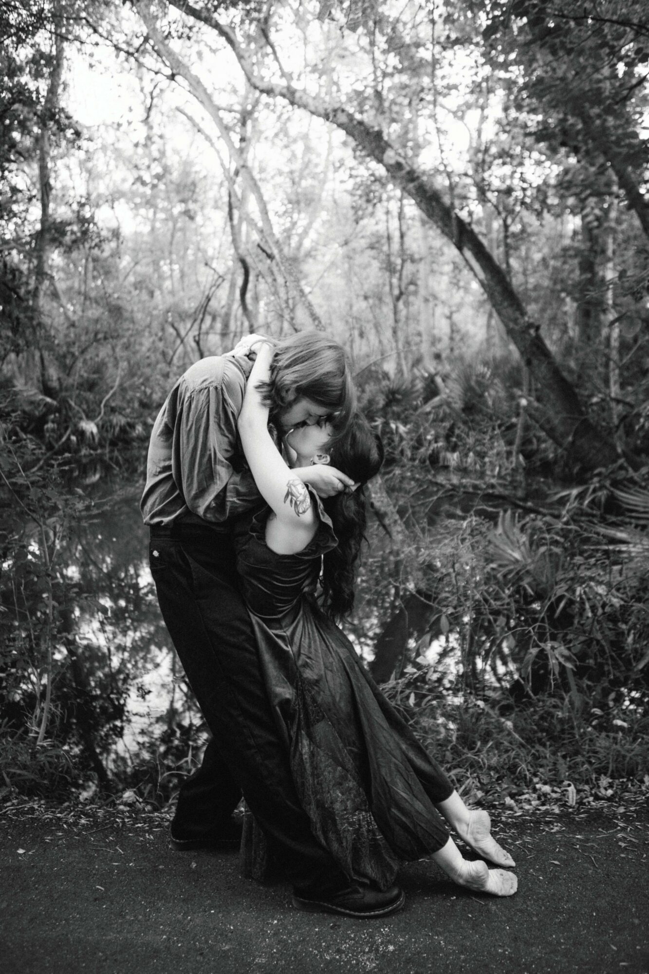 A couple dancing in a forest near a body of water, with trees and foliage in the background.
