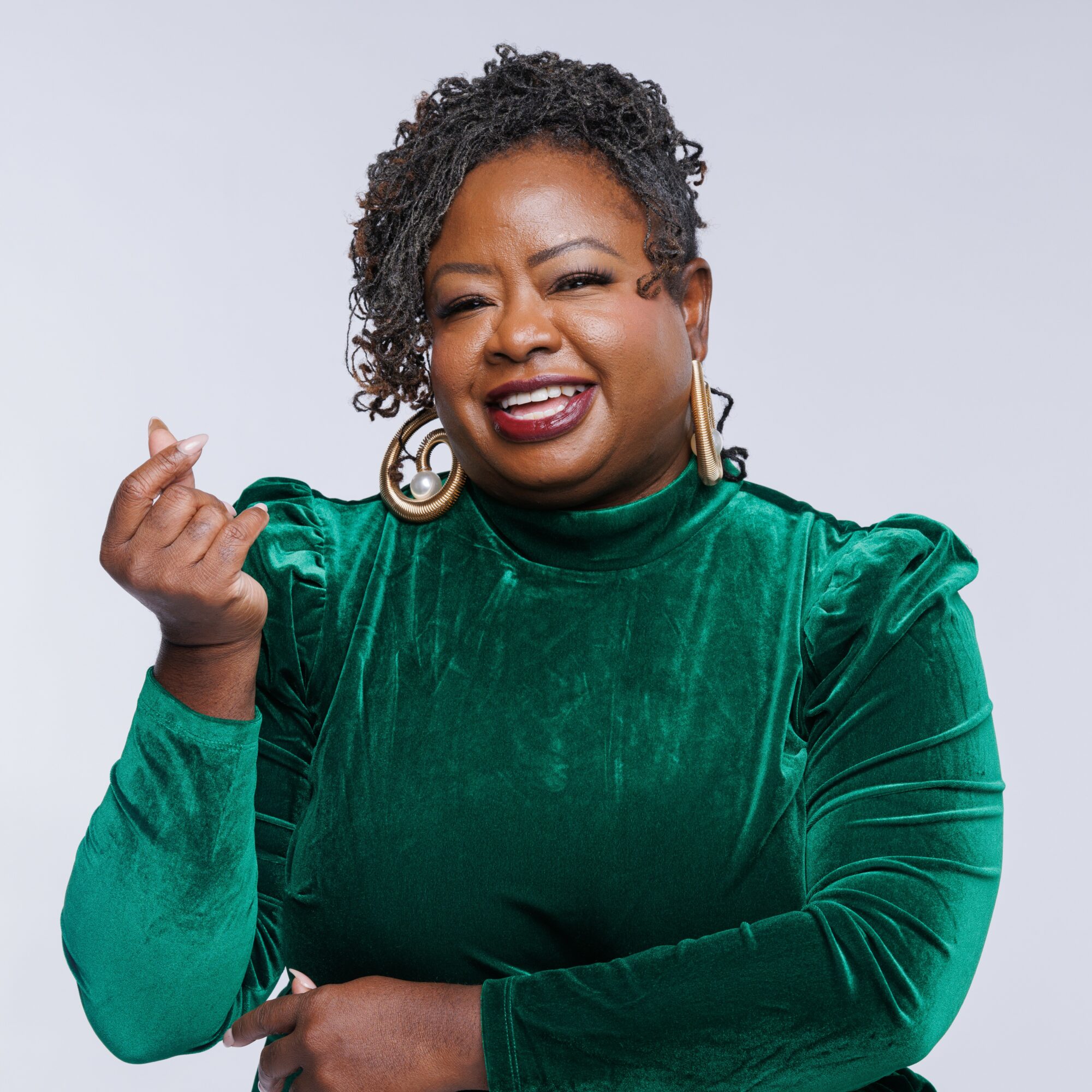 Woman with short curly hair wearing a green velvet top, smiling, making a hand gesture, against a light background.