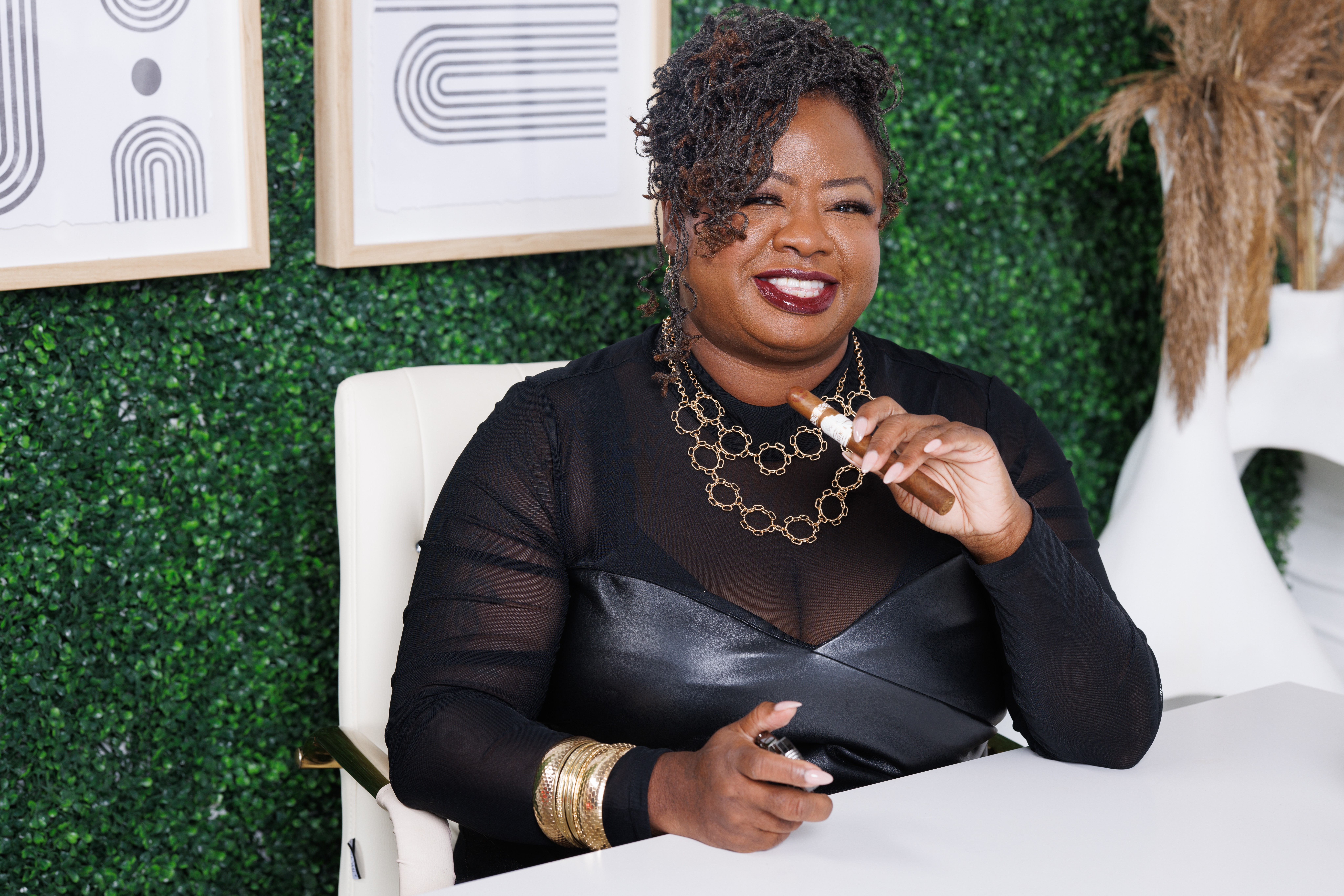 Smiling woman with short curly hair wearing black top and gold jewelry, sitting in a white chair.