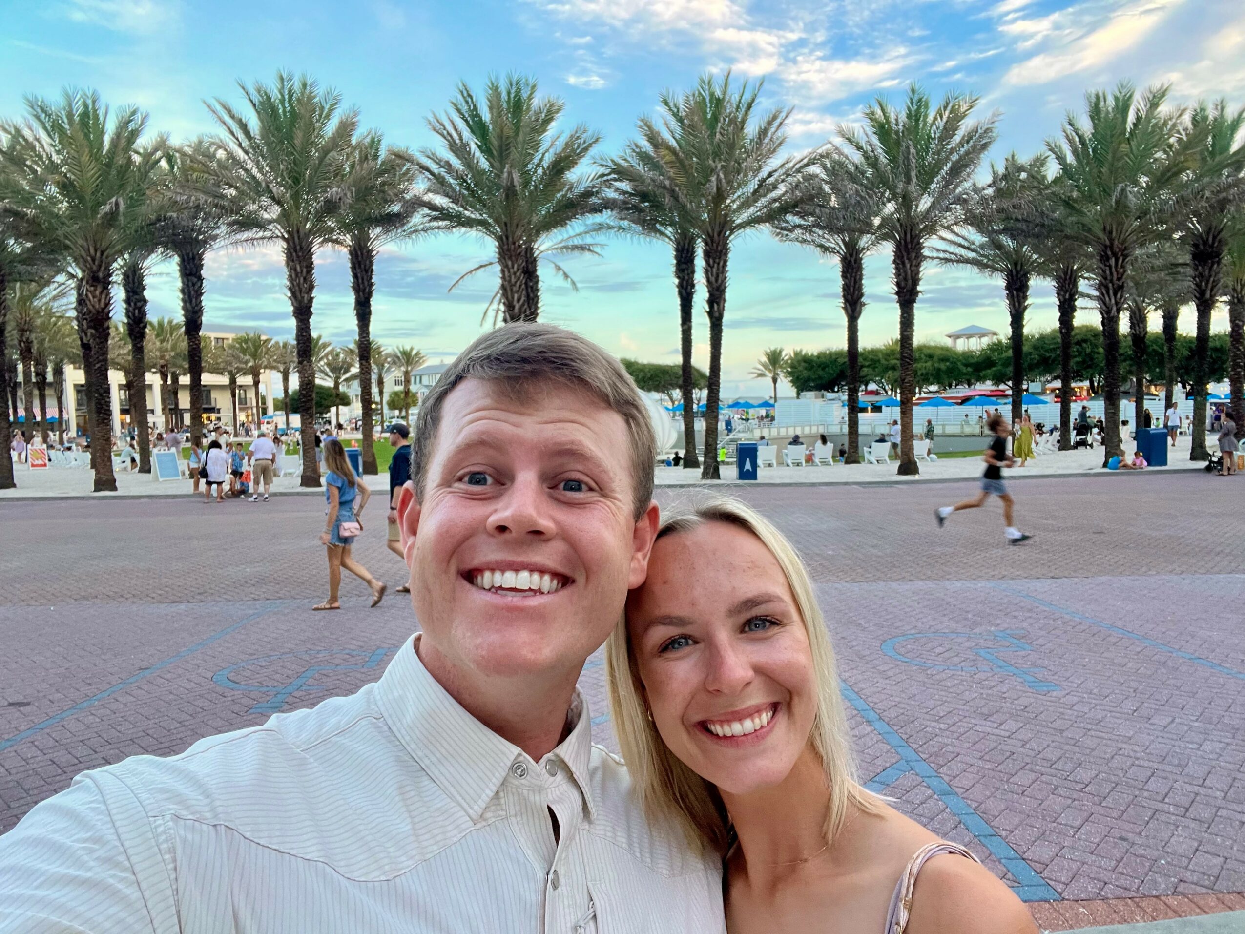 Two smiling people taking a selfie in front of palm trees and a waterfront with boats and people walking.