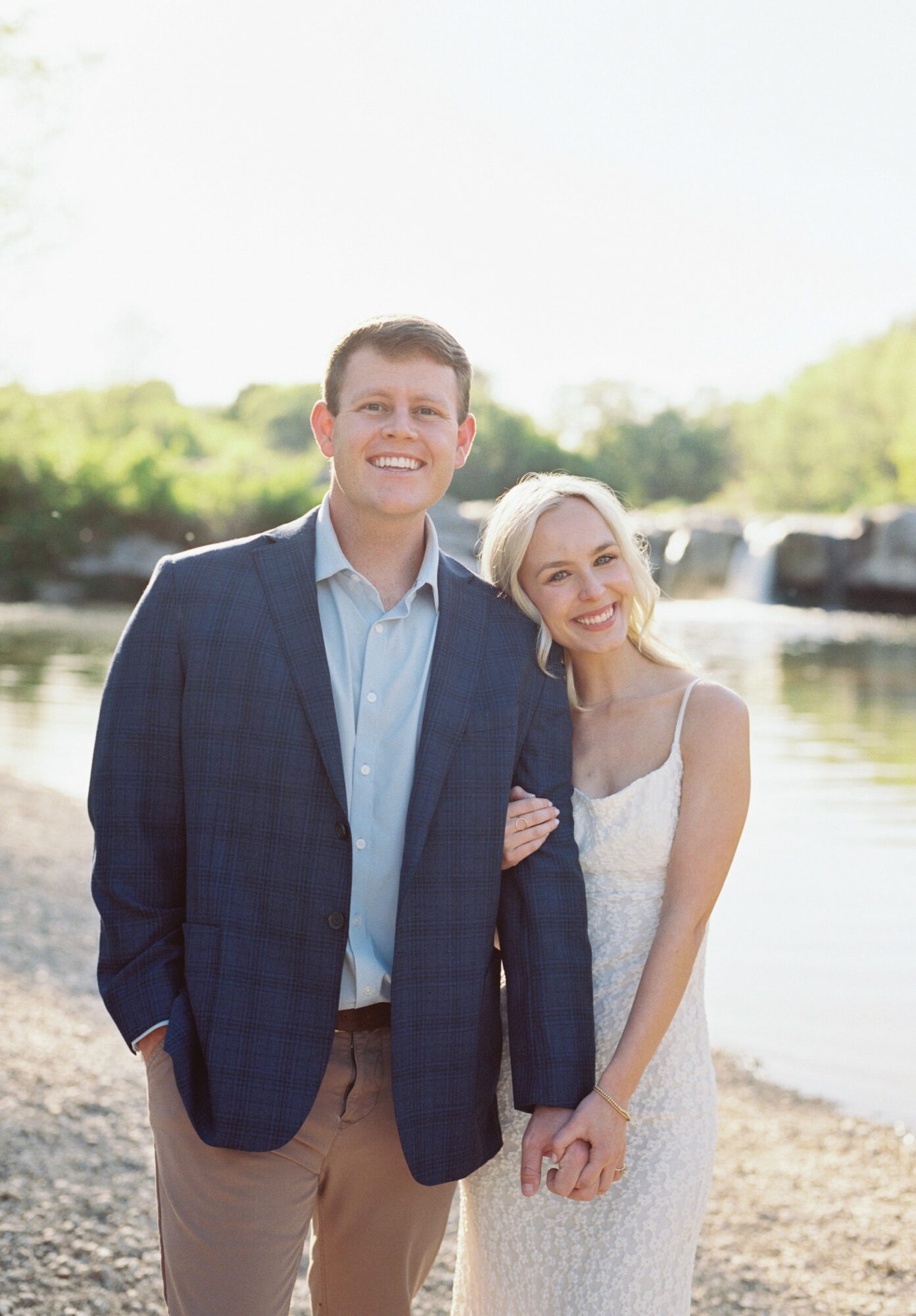 A smiling couple standing outdoors near water, with trees and a bridge in the background.