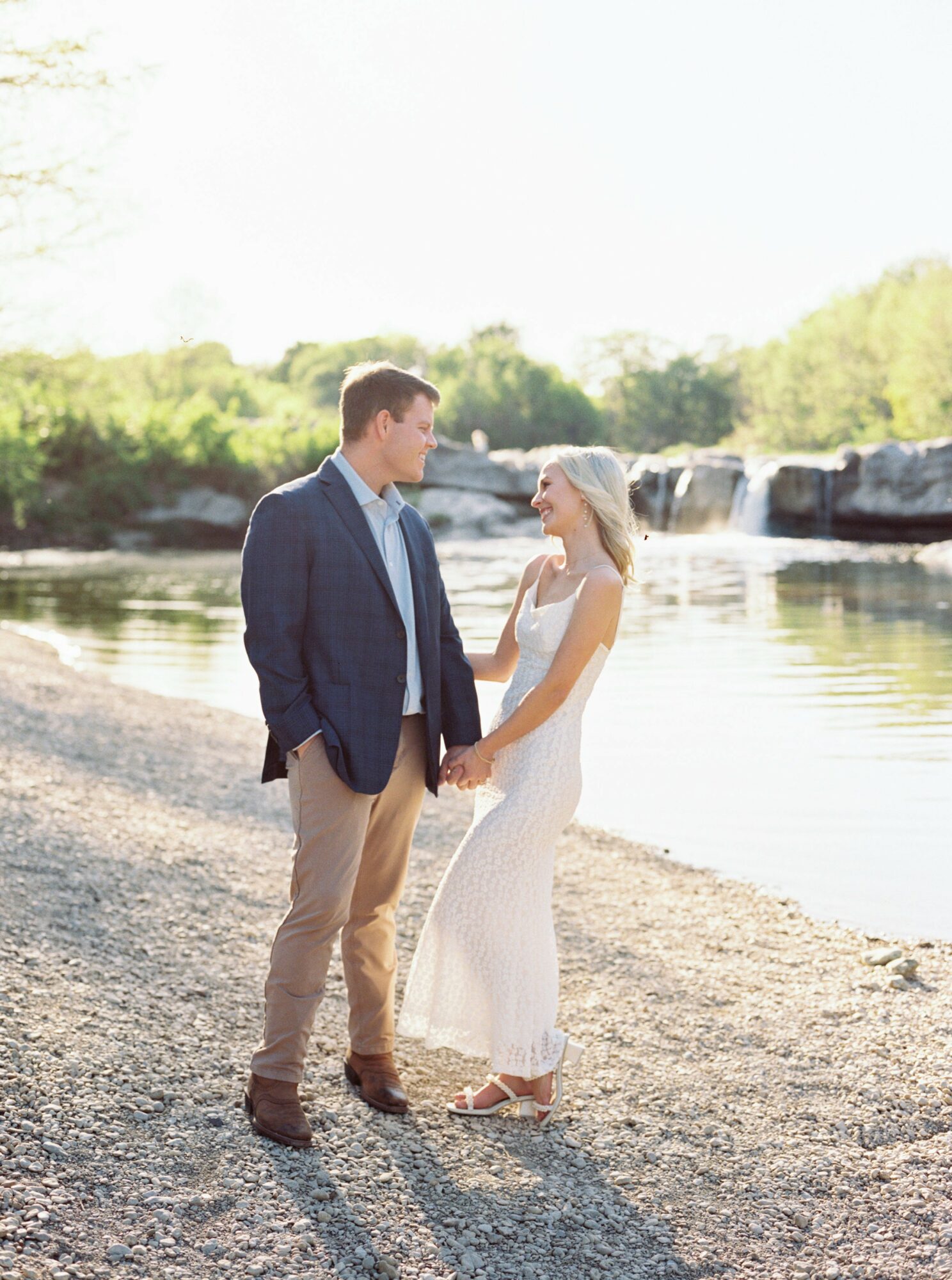 A couple holding hands by a river with a waterfall in the background, standing on a rocky shore.