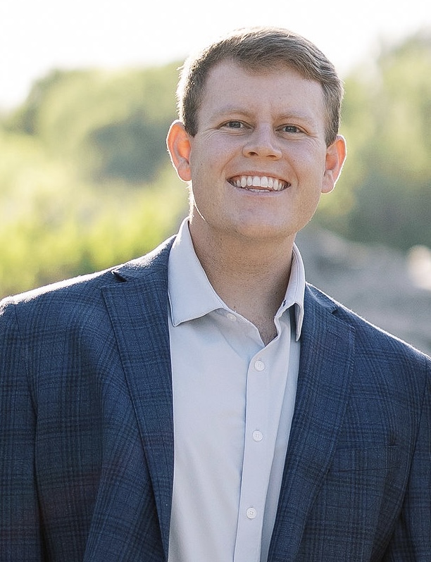 Young man smiling outdoors, wearing a suit jacket and white shirt, with blurred greenery background.