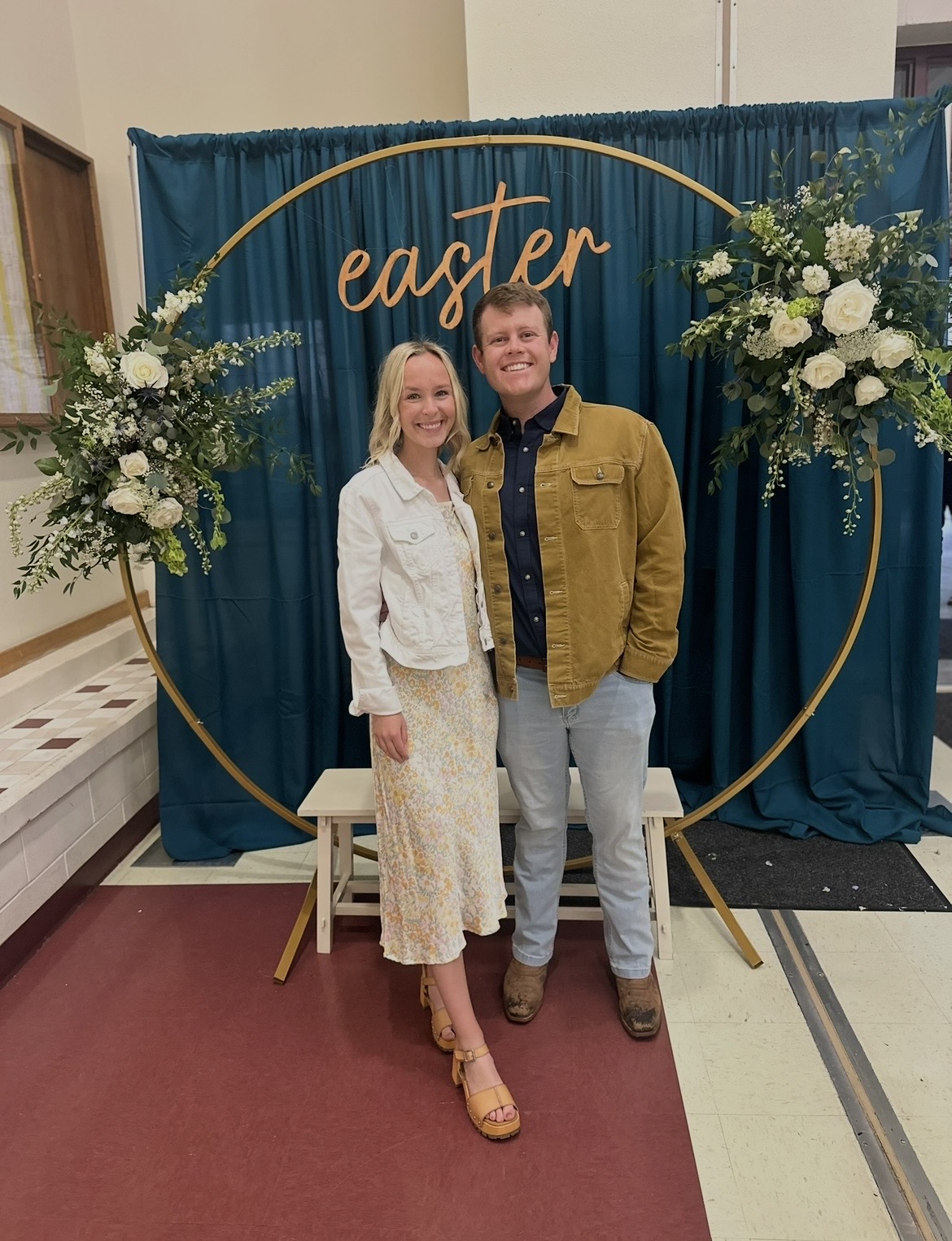 A smiling couple stands in front of a blue backdrop with gold lettering and floral arrangements, celebrating Easter.