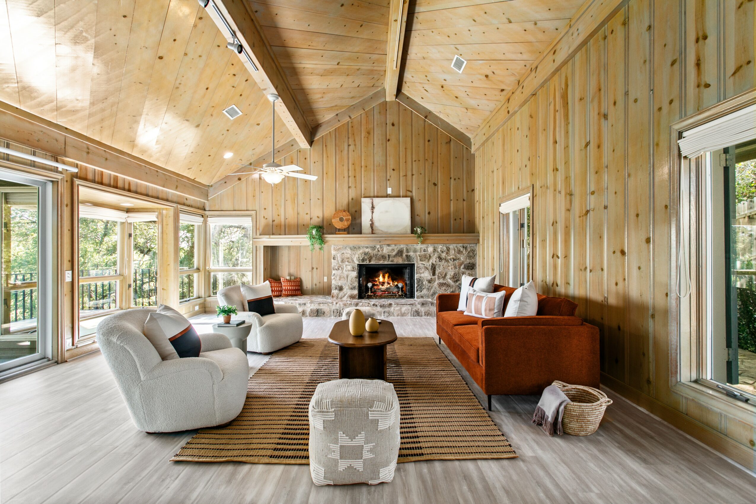 Living room with wooden walls and ceiling, fireplace, large windows, and various chairs and sofas around a rug.