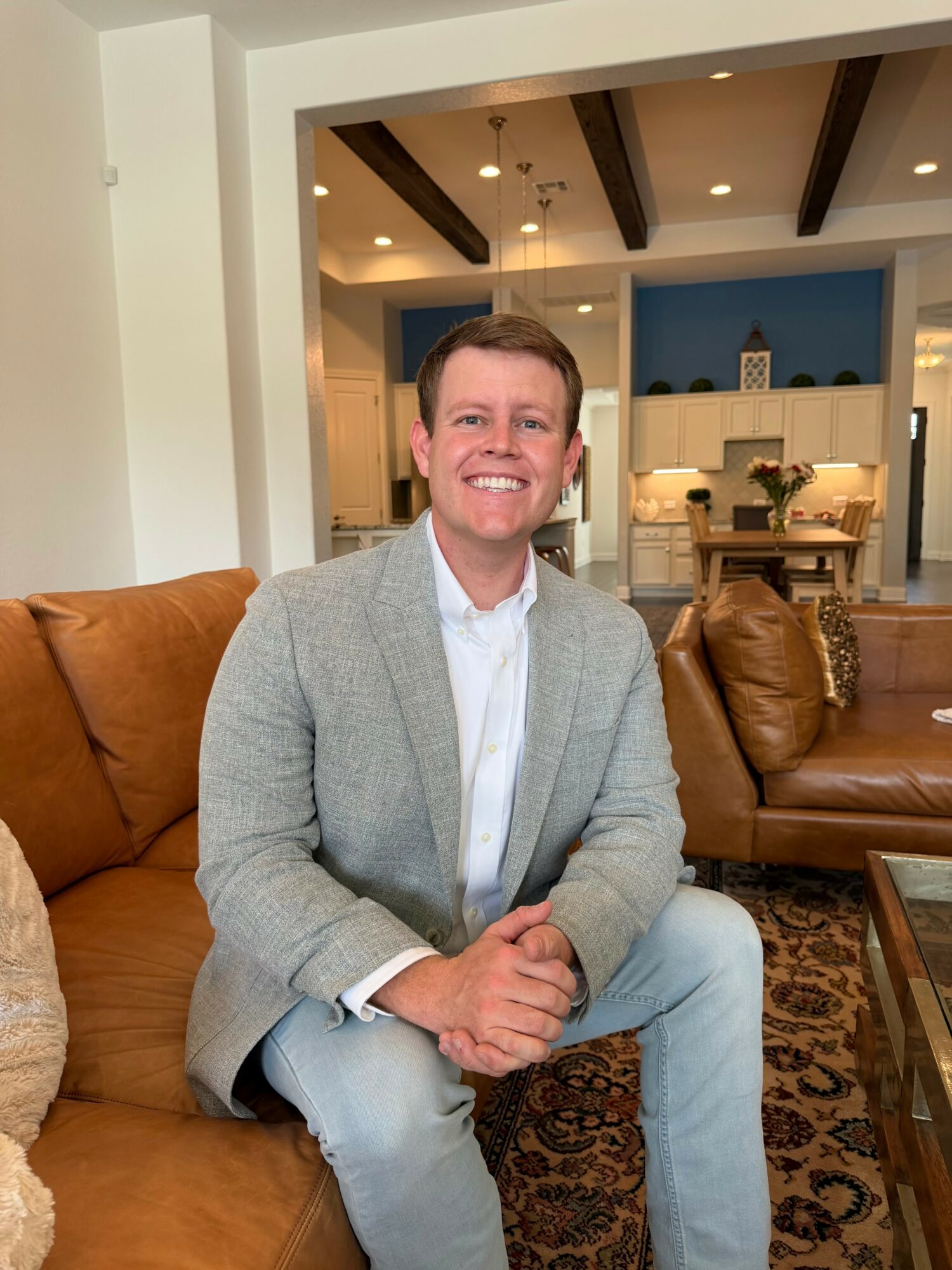 Man sitting on a sofa, smiling, in a living room with modern decor and wooden ceiling beams.