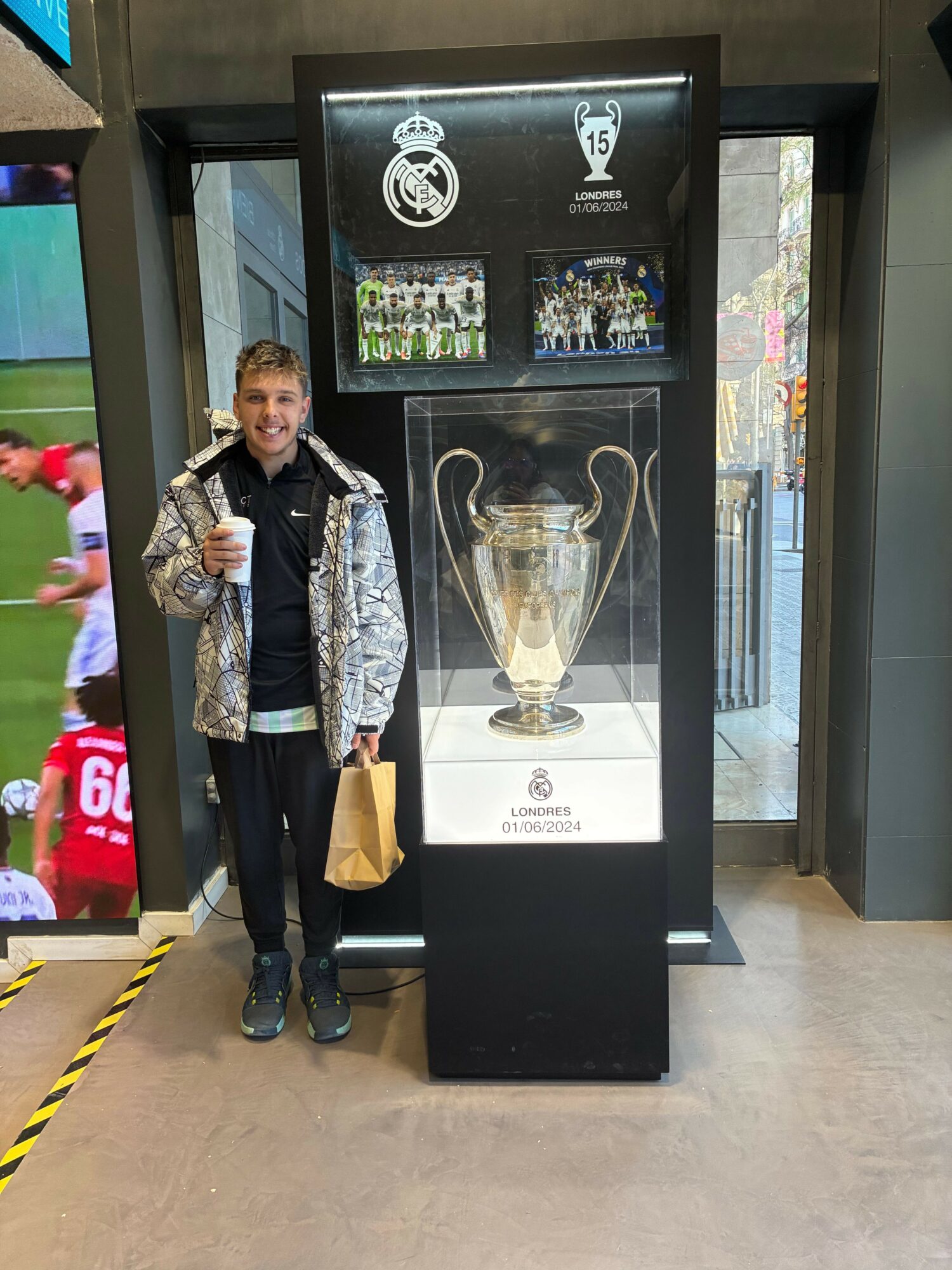 Young man standing next to a display case with a trophy, holding a drink and a bag, smiling.