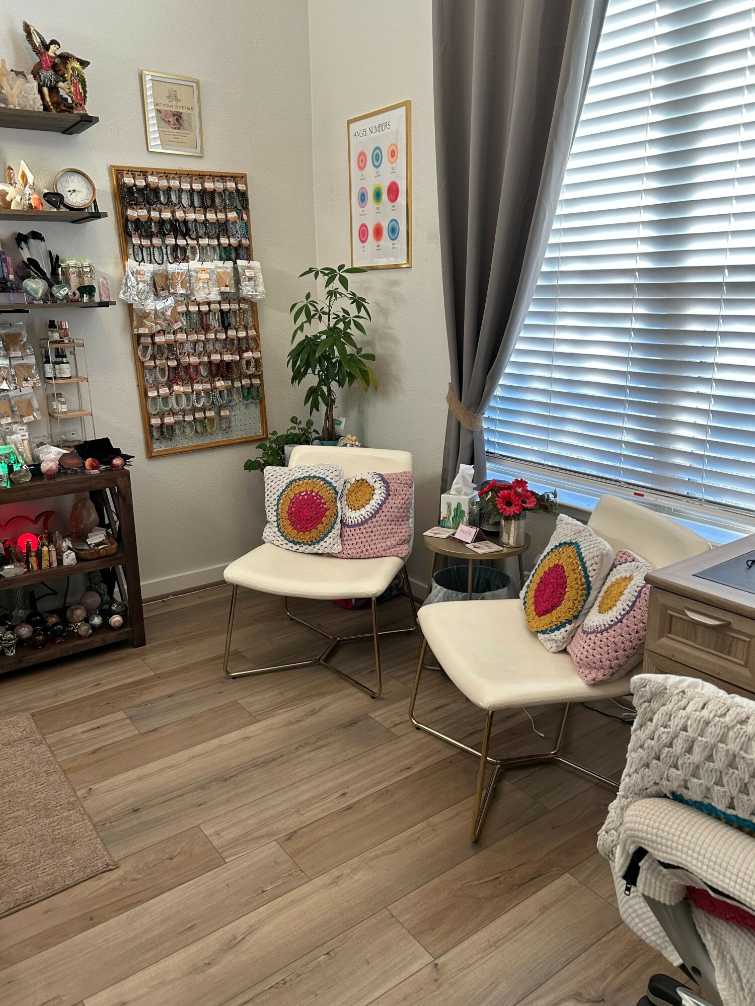 Two white chairs with colorful circular pillows in a cozy room with shelves, plants, and window with blinds.