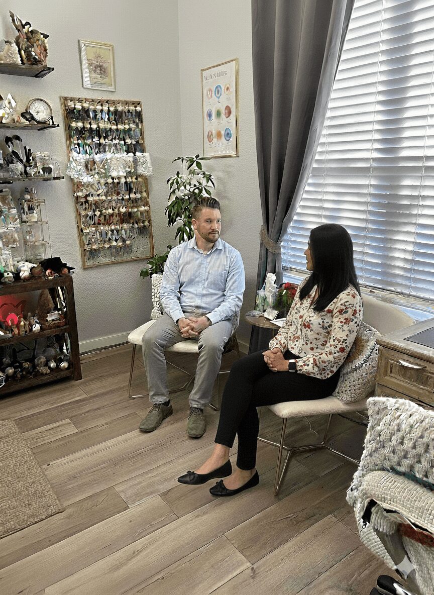 Two people sitting and talking in a room with shelves, plants, and window with curtains.