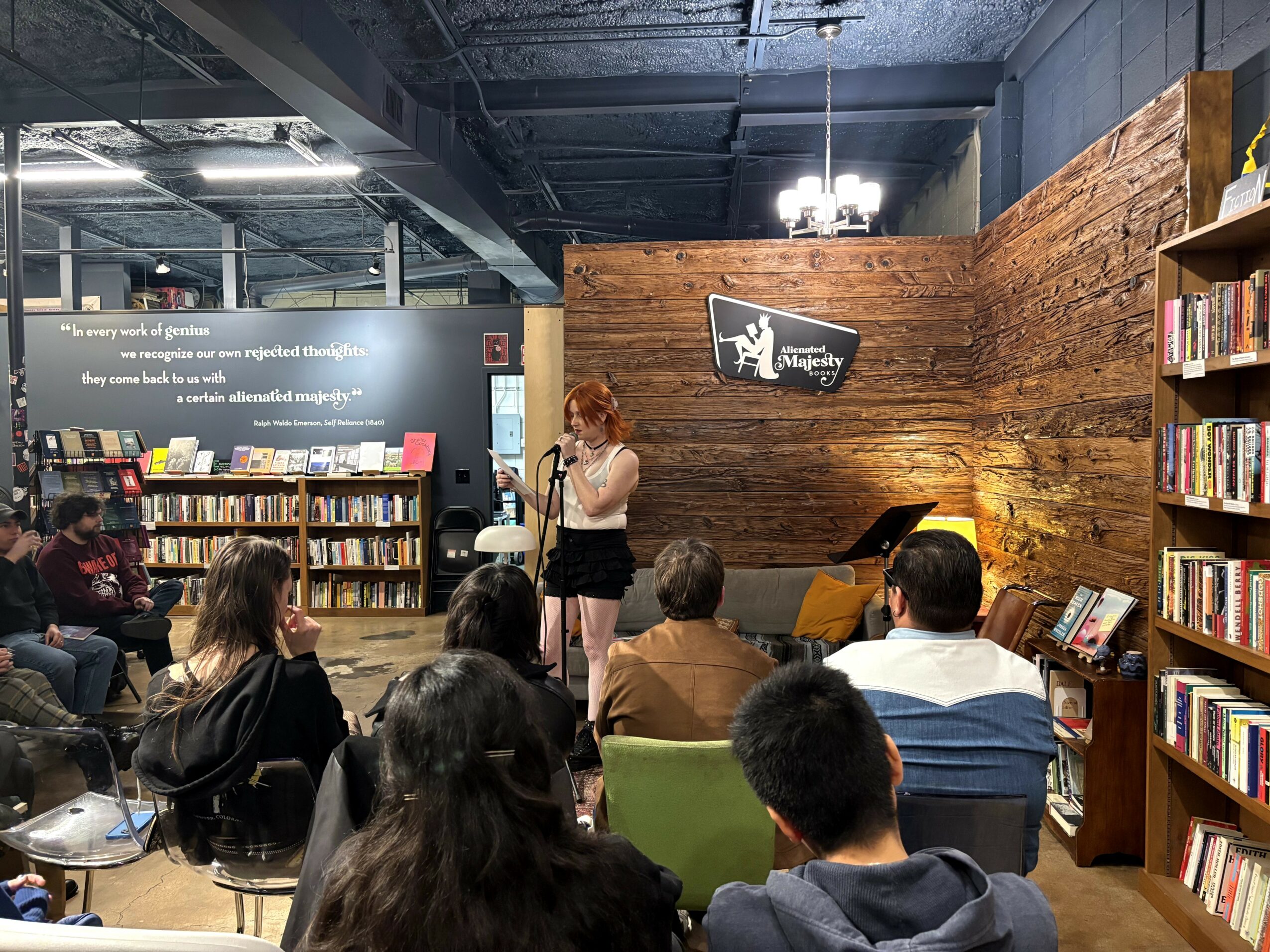 Woman speaking to an audience in a bookstore or library with bookshelves and a wooden wall behind her.