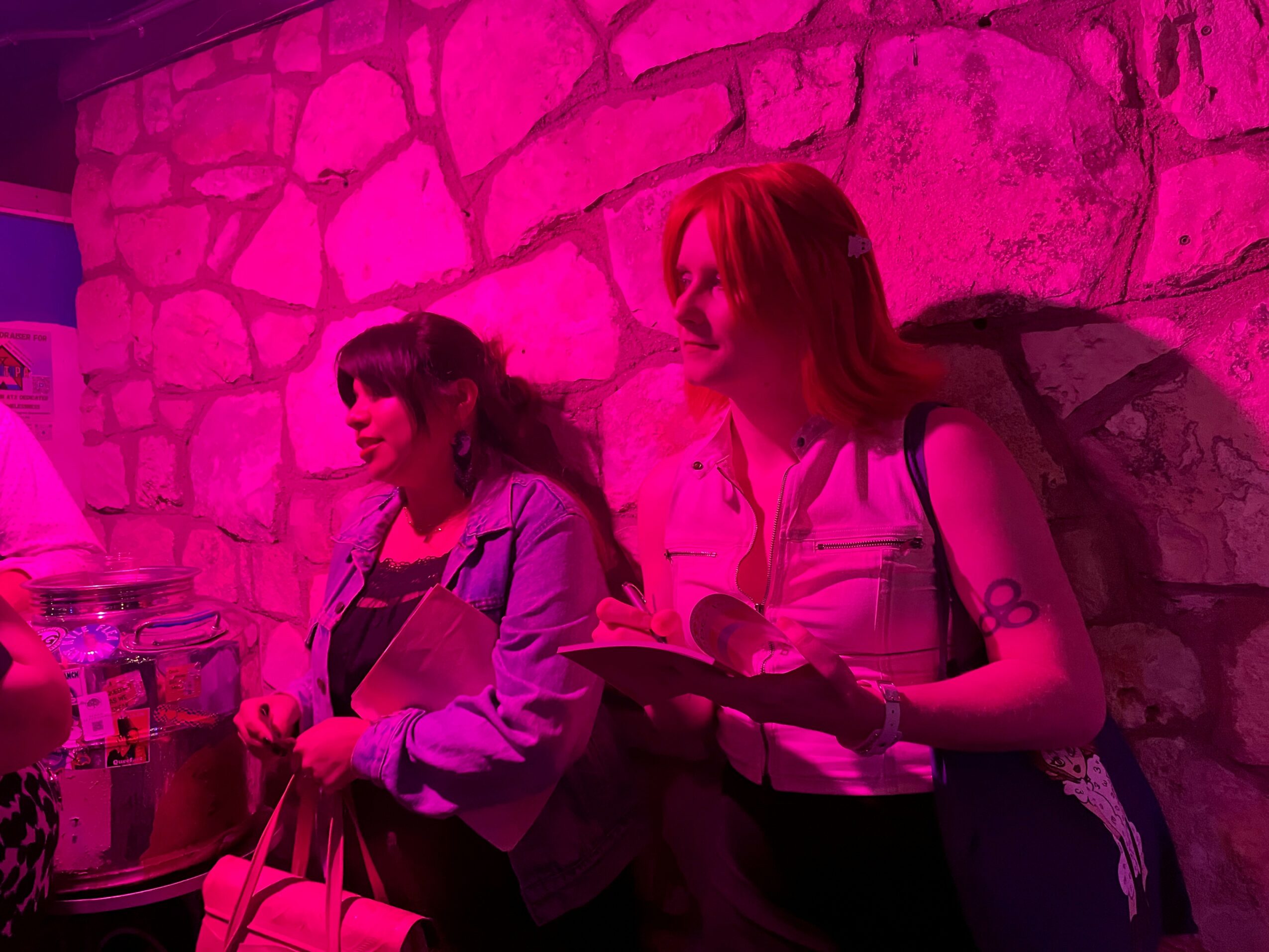 Two women stand against a pink-lit stone wall, one holding a notebook and pen, the other with a bag.
