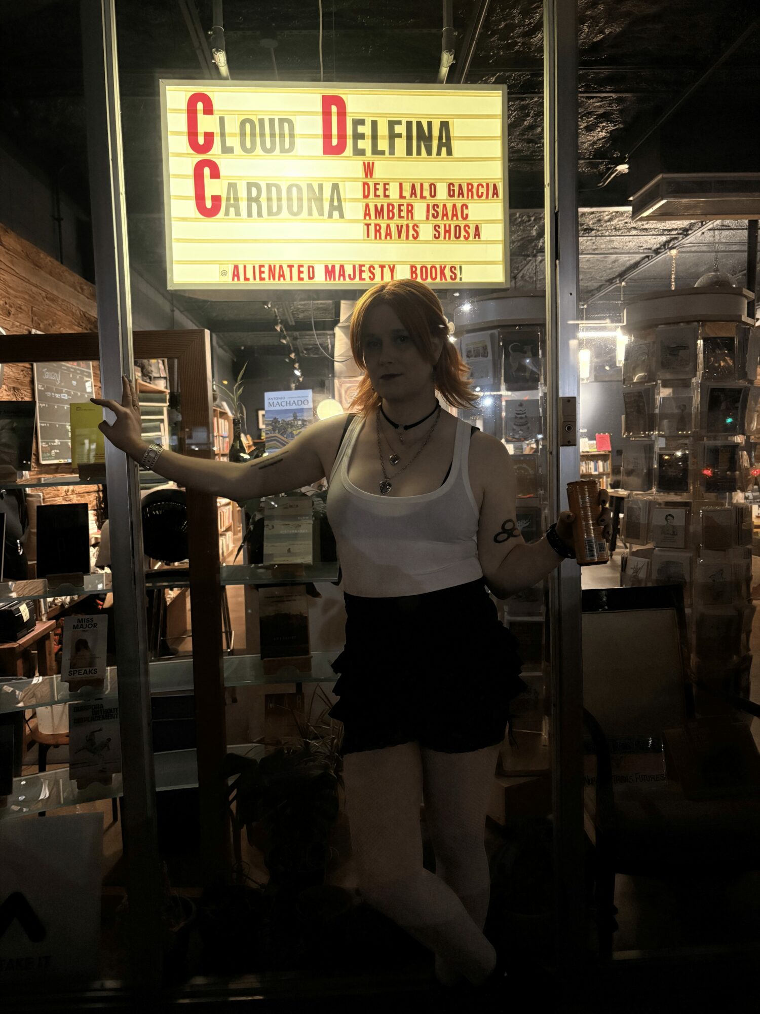 Woman with curly hair in white top and black shorts standing inside a dimly lit venue, holding a drink.